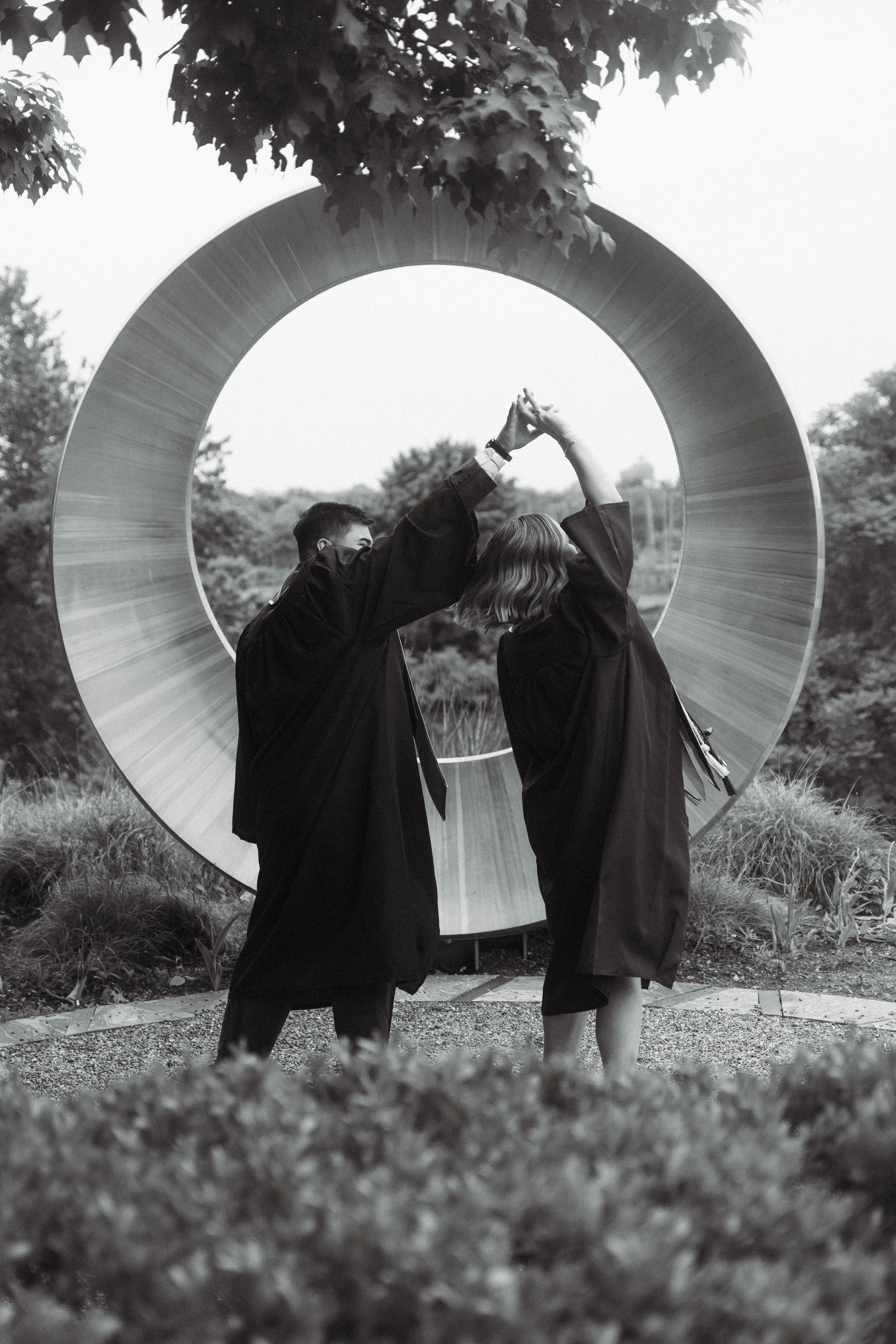 Two graduates in robes are holding hands up in celebration in front of a large, modern circular sculpture outdoors.