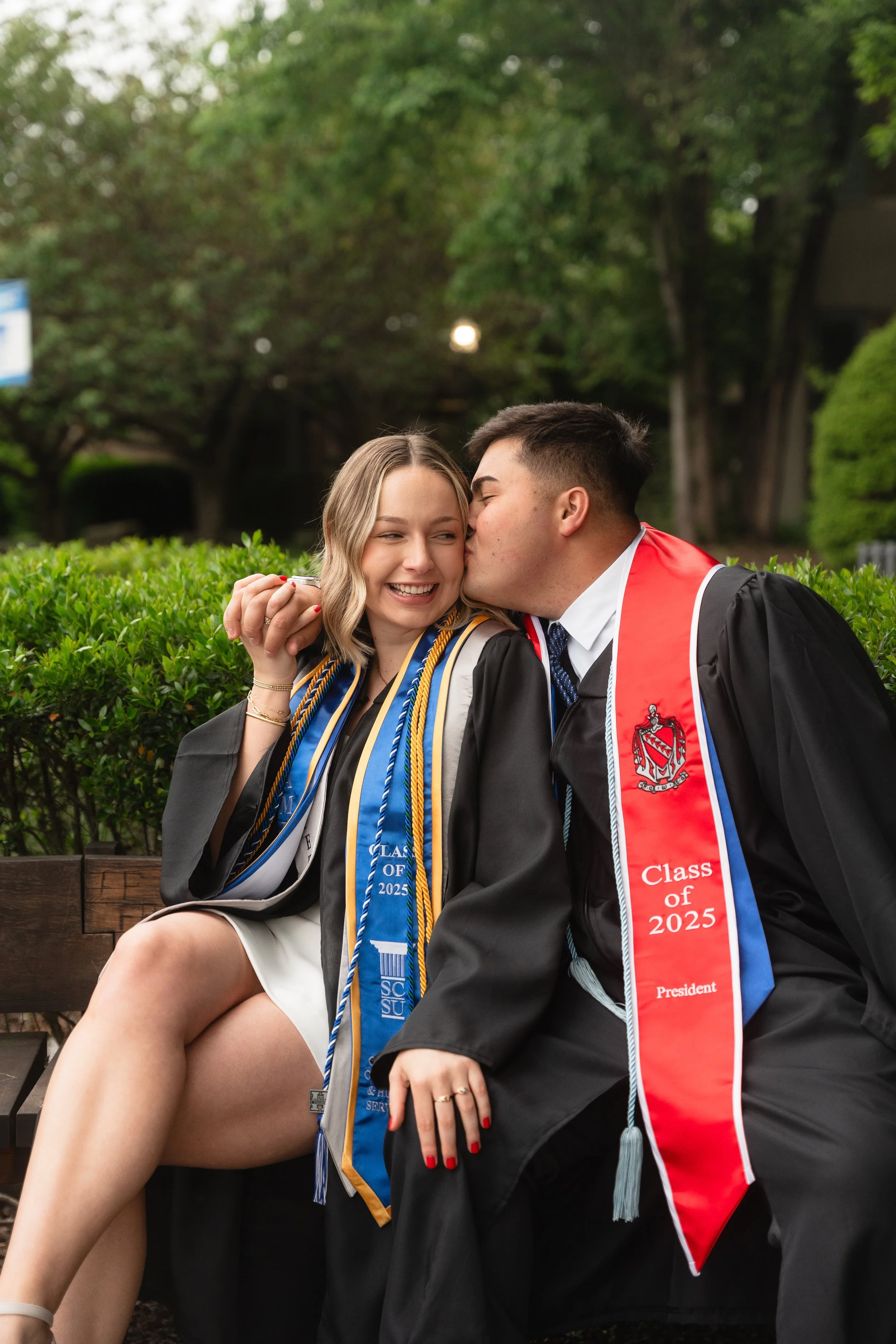 Two graduates in caps and gowns sitting on a bench, smiling and sharing a kiss. The woman is wearing several honor cords, and the man is kissing her on the cheek.