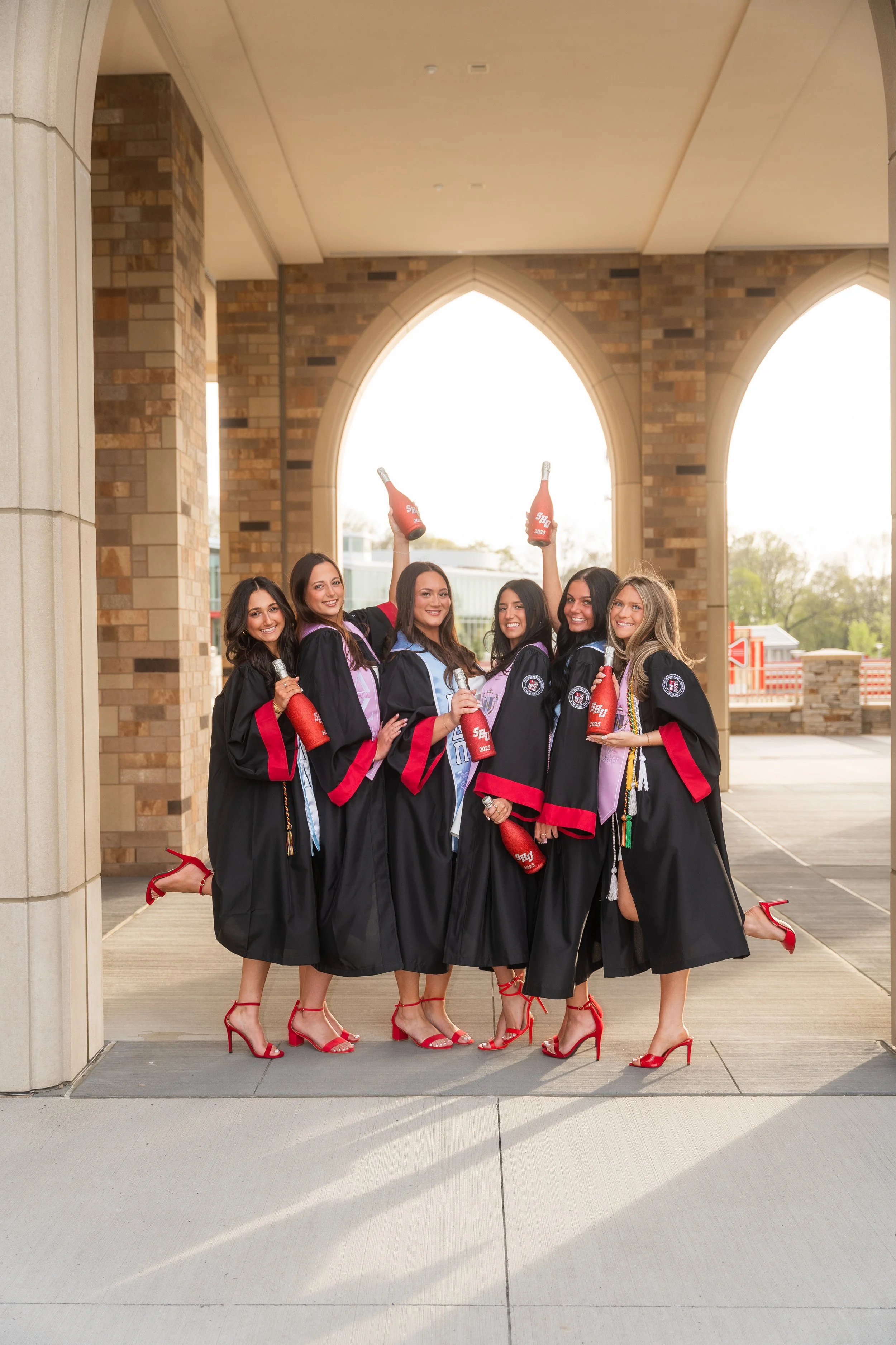 Group of six women in graduation gowns and red high heels celebrating outdoors under archways, holding bottles of sparkling beverage.