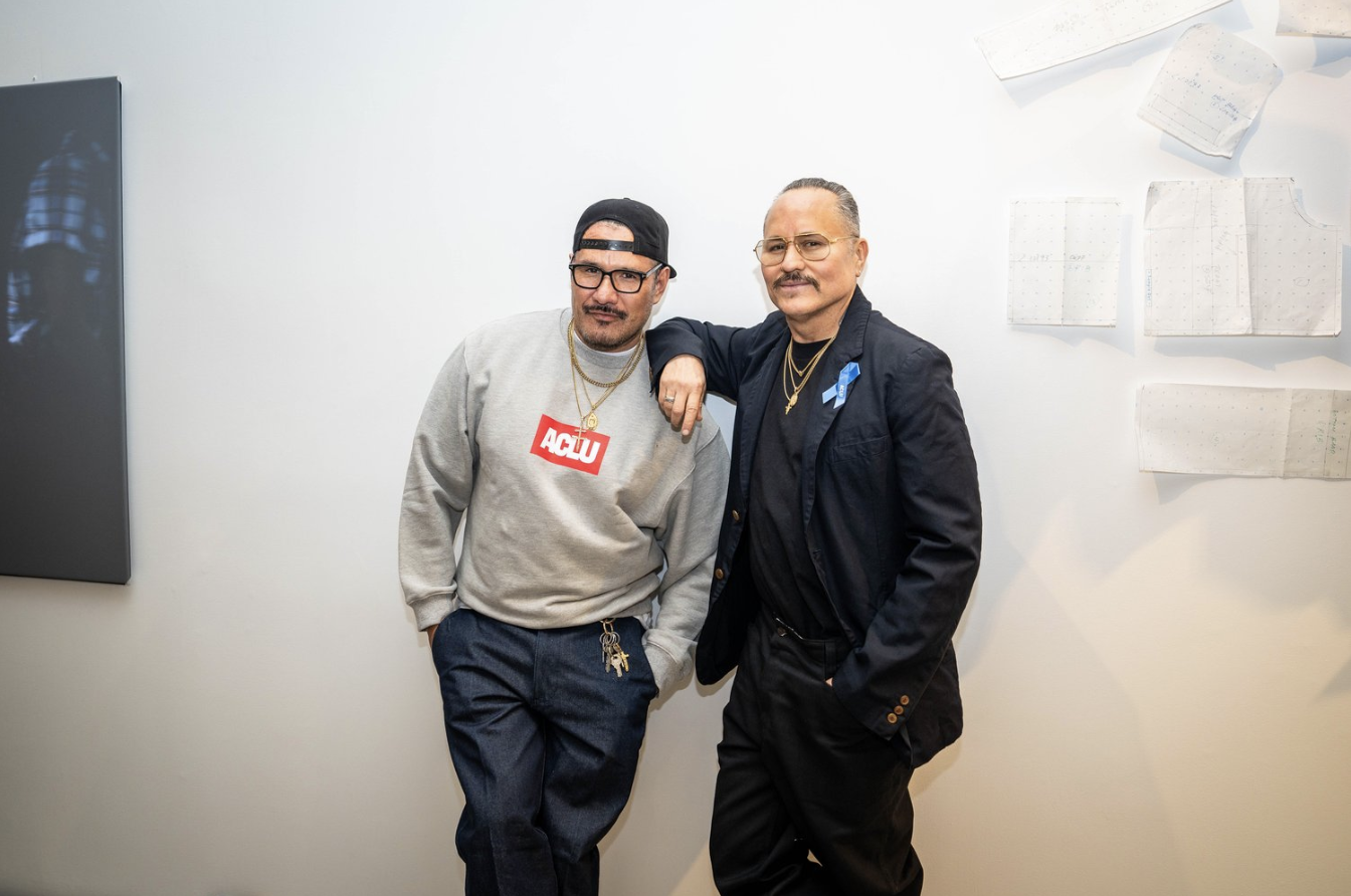 Willy Chavarria (right) leans against a white wall in a black suit with gold necklaces and a blue ribbon. Beside him, a man in a gray ACLU sweatshirt and jeans stands casually. Both look at the camera