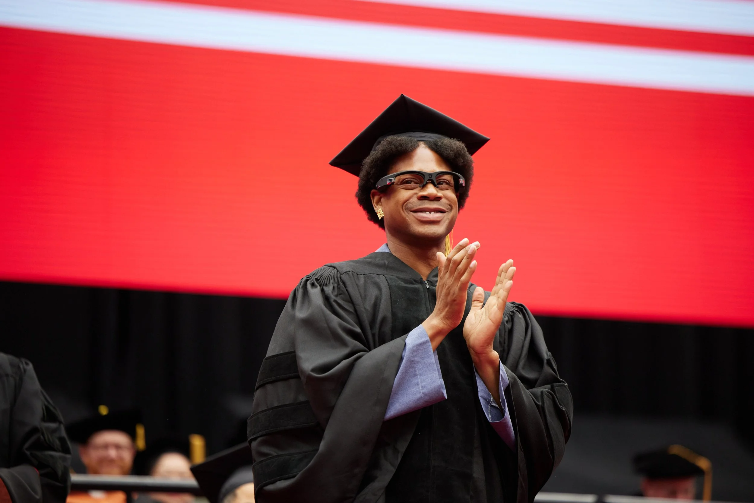 Fashion designer Telfar Clemens, wearing a black graduation cap and gown with blue satin lining, claps while smiling during The New School graduation ceremony. A red backdrop contrasts with his outfit.