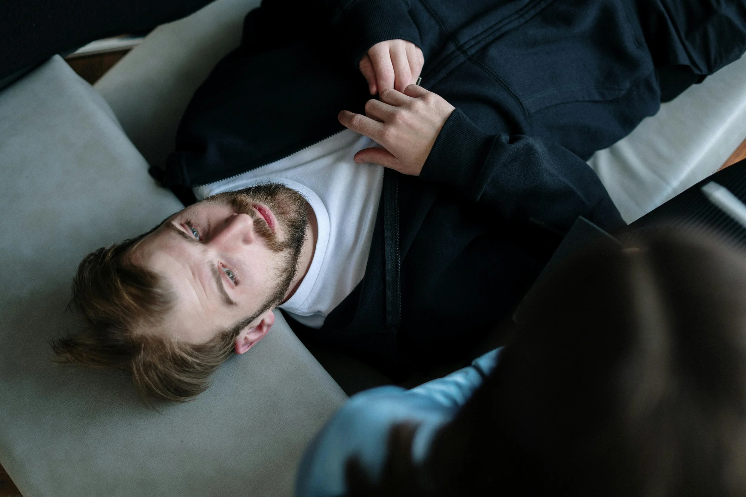 En mannlig person ligger på en grå sofa og reflekterer over tankene sine i et terapeutisk miljø. Fotografi: Arkivfoto fra Cottonbro.