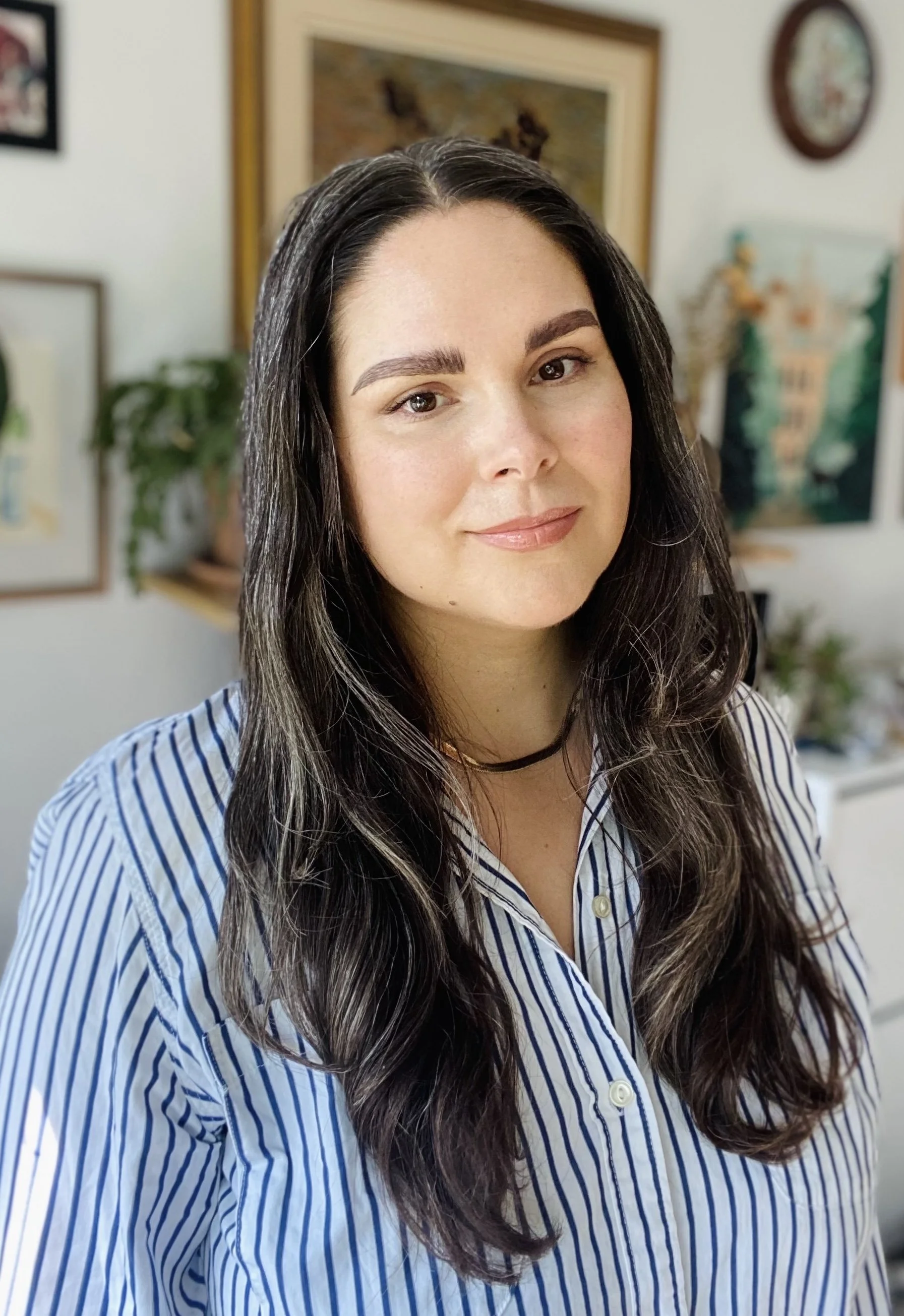 Roberta Hayes, FMHC at FMU in Waterloo woman with long dark hair, wearing a striped blue and white shirt, smiling softly. Background includes framed artwork and indoor plants.