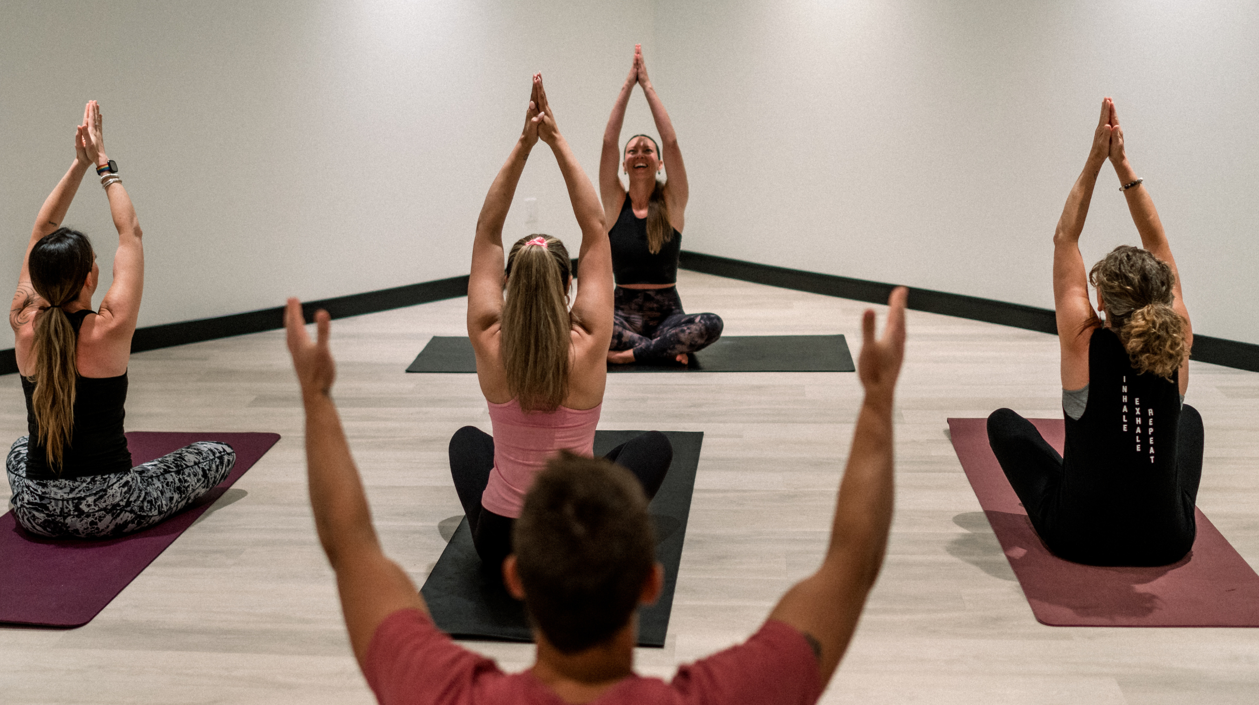 Yoga class with five women seated on yoga mats in a cross-legged position, raising their arms and hands in the air, with a smiling instructor leading the session in a bright, plain room.