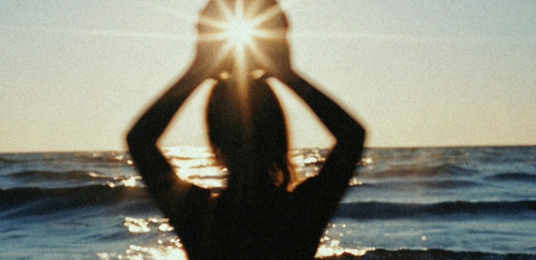 Woman at the water at sunset with arms raised, calm and looking outward.