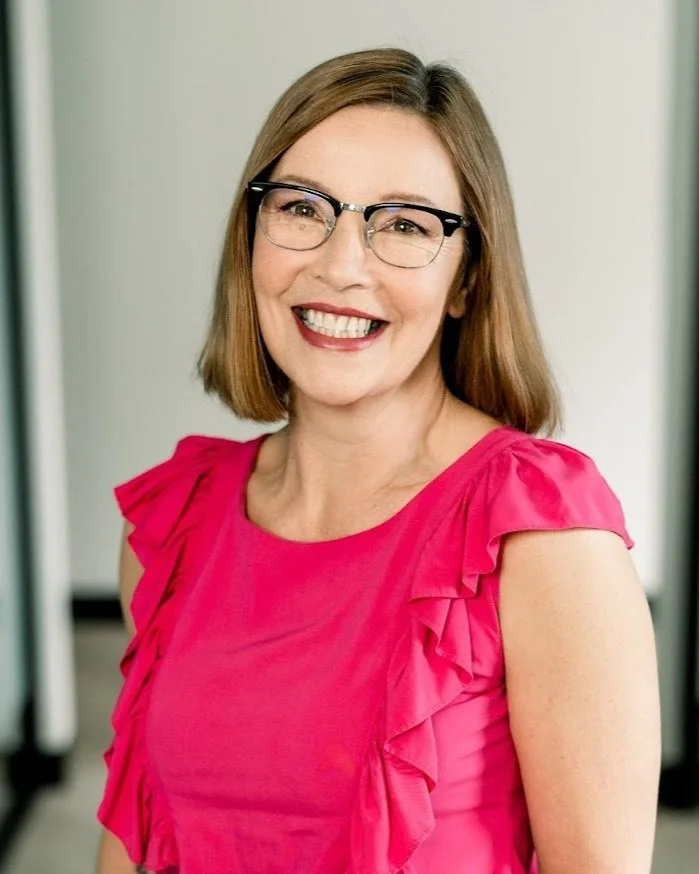 Gloria Vaughan, FMHC at Functional Medicine Uptown in Waterloo smiling woman wearing glasses and a pink ruffled top, standing indoors.