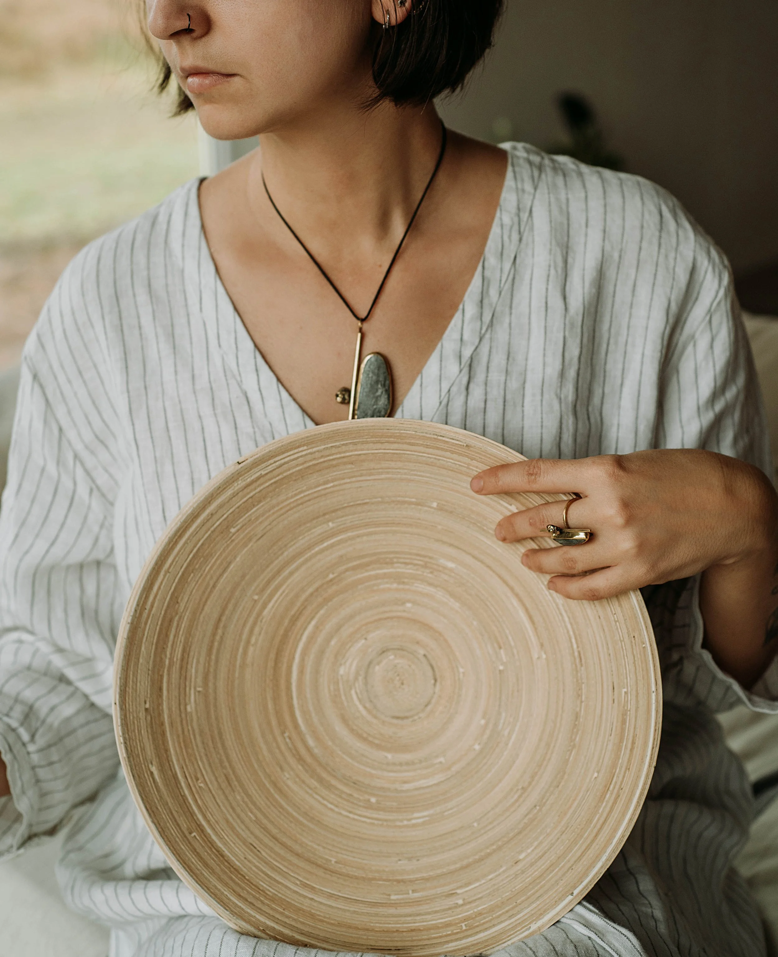 A woman wearing a striped white shirt holding a round, beige bamboo plate with concentric circles, wearing a stone pendant