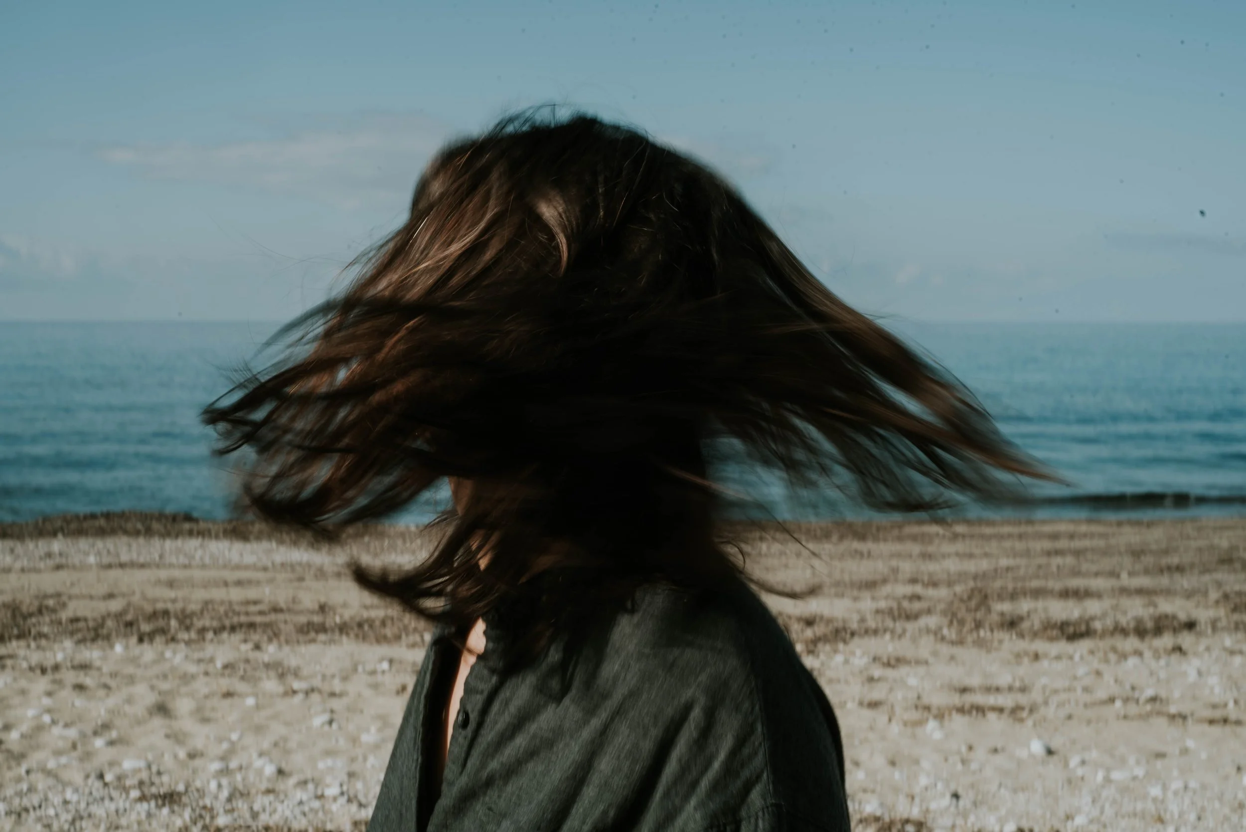 Person with medium-length hair blowing in the wind on a beach with the ocean in the background.