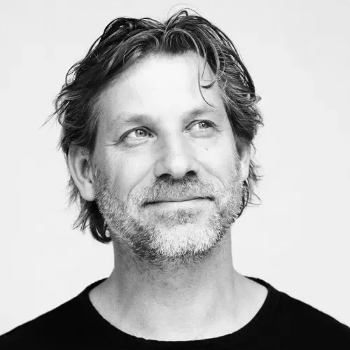 A black and white portrait of a middle-aged man with medium-length wavy hair and a beard, looking slightly upward and to the side, wearing a dark shirt against a plain background.