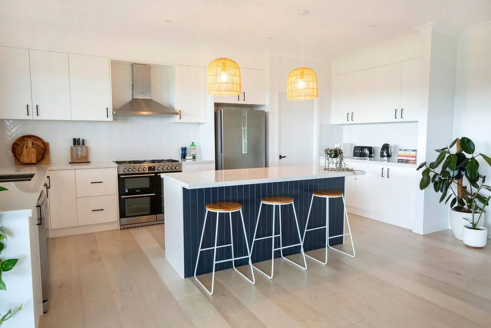 Modern kitchen with white cabinets, a blue island with three stools, stainless steel appliances, and potted plants.