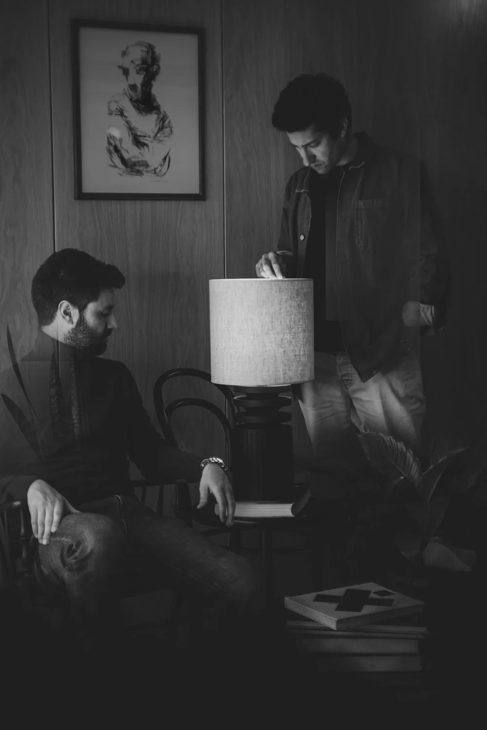 Two designers are portrayed in a living room with wooden walls; one sitting and one standing, near a table lamp on a side table with books. A framed photo or art hangs on the wall.