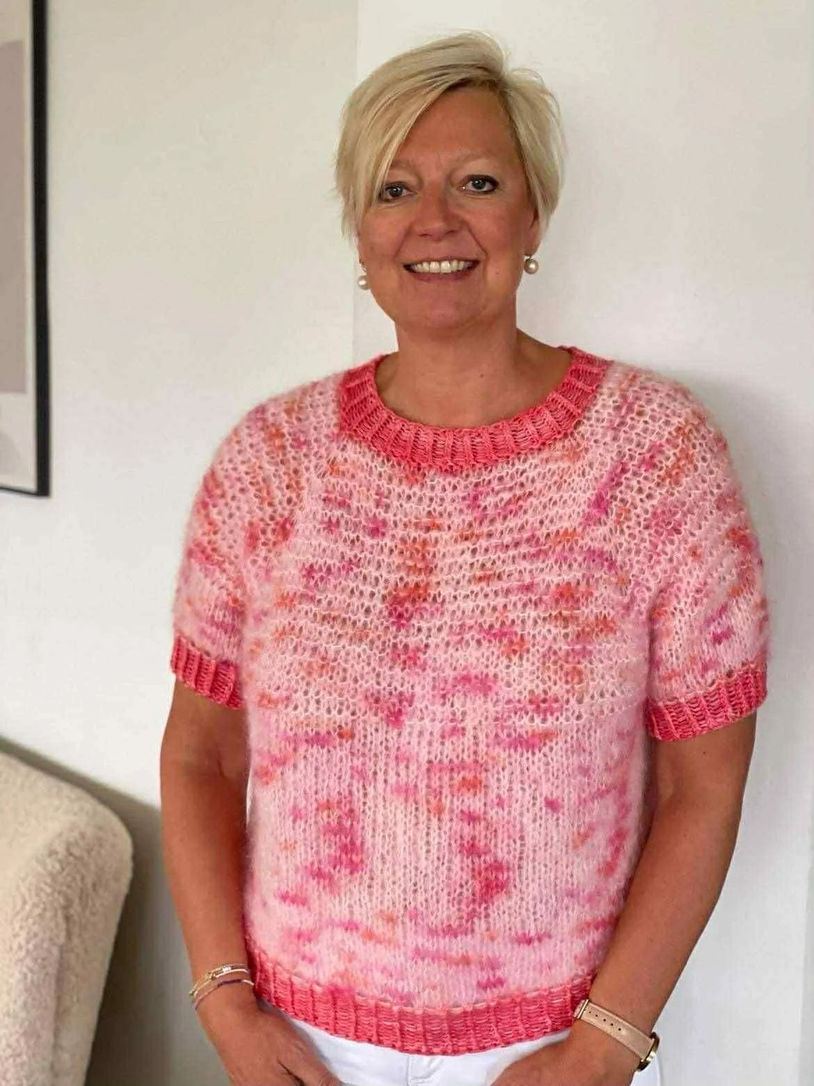 Kvinde med kort blond hår, smilende, iført lyserød og hvid strikket sweater, stående indendørs mod en hvid væg.