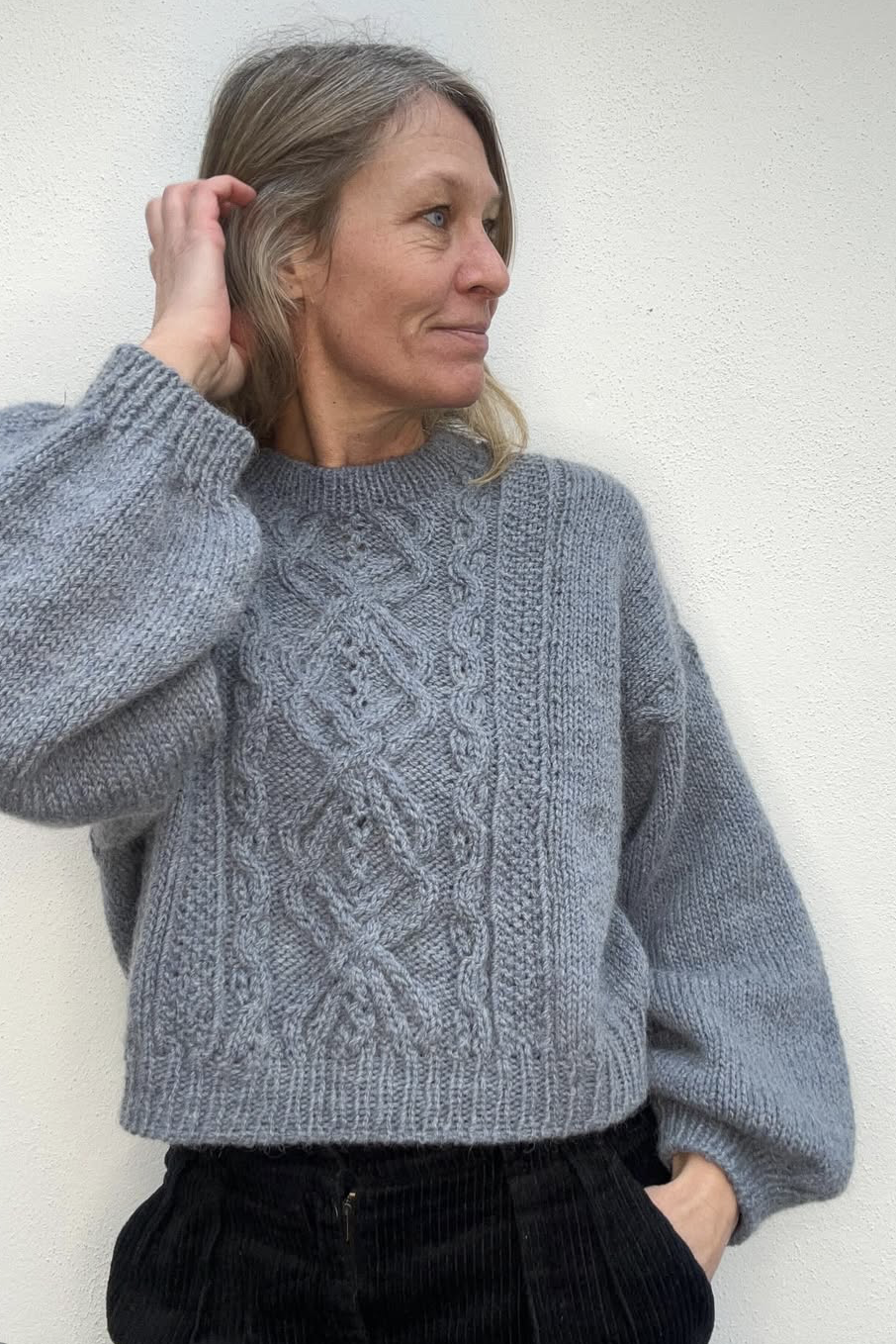 Kvinde med blond hår og en grå strikketrøje med kabelmønster, der står mod en hvid væg.