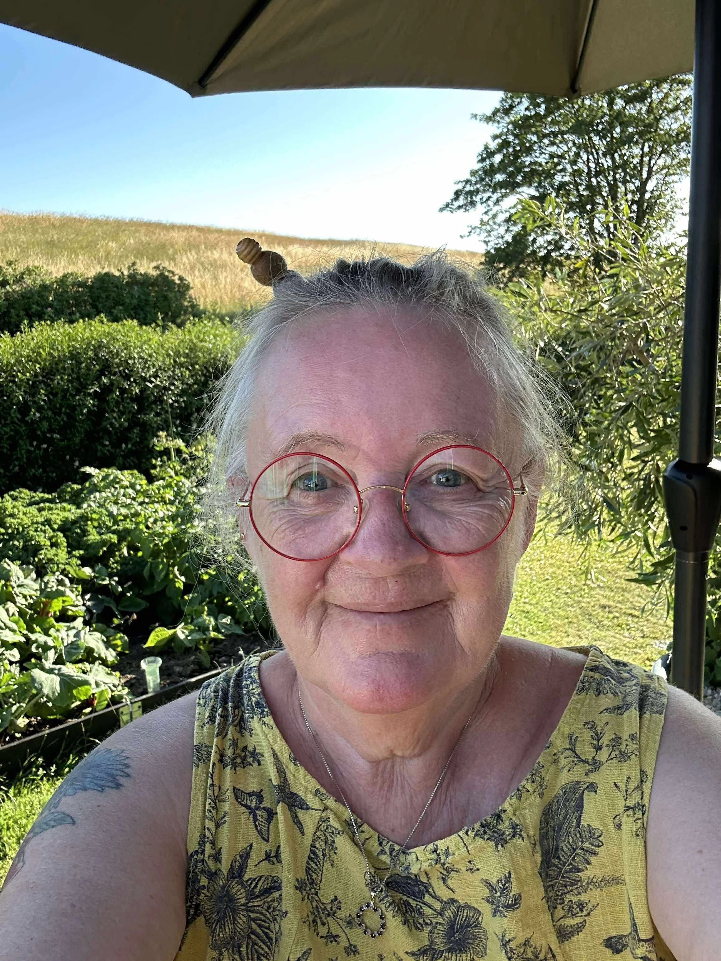 Et portræt af en ældre kvinde med brunt hår i en beskåret frisure, iført store røde glas, en gul blomstret top, og en halskæde med et hjertefedt vedhæng. Hun står under en parasol i en have med grønne buske og træer i baggrunden på en solrig dag.