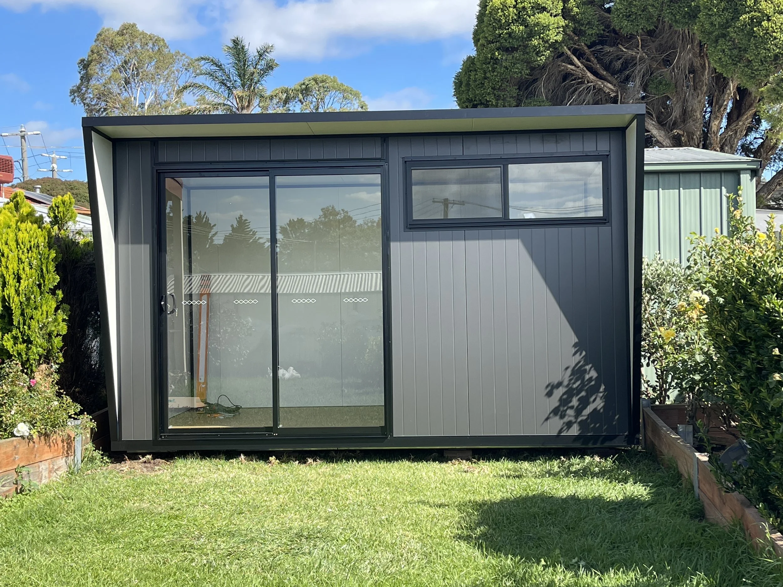 Modern backyard pod in Monument colour with VJ groove cladding, cantilever roof design, and large sliding glass door, installed in a landscaped Melbourne garden.