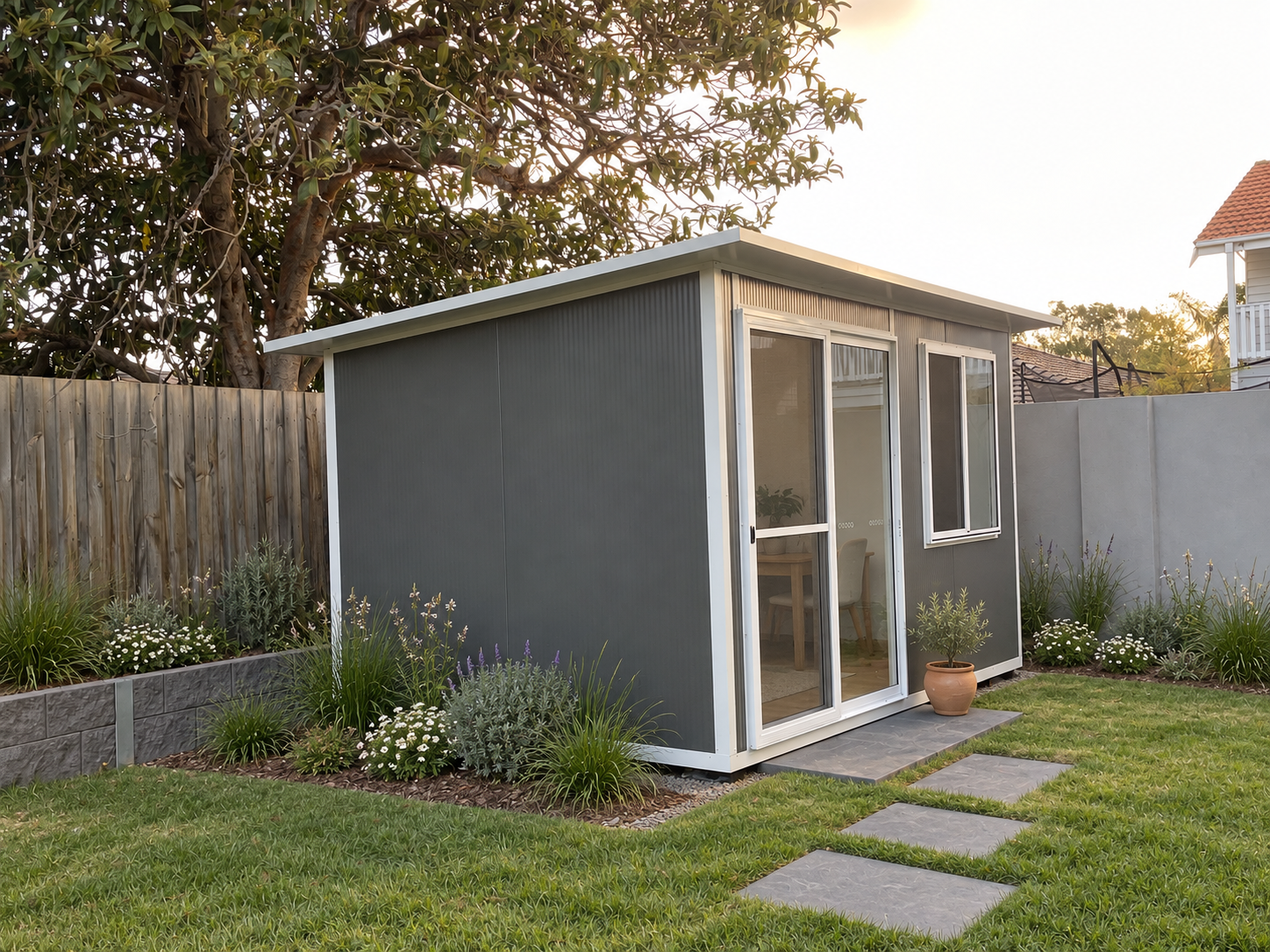 A small gray backyard shed with glass sliding doors and a window, surrounded by a lawn, stepping stones, and flower beds with plants and white flowers, enclosed by wooden and concrete fences.