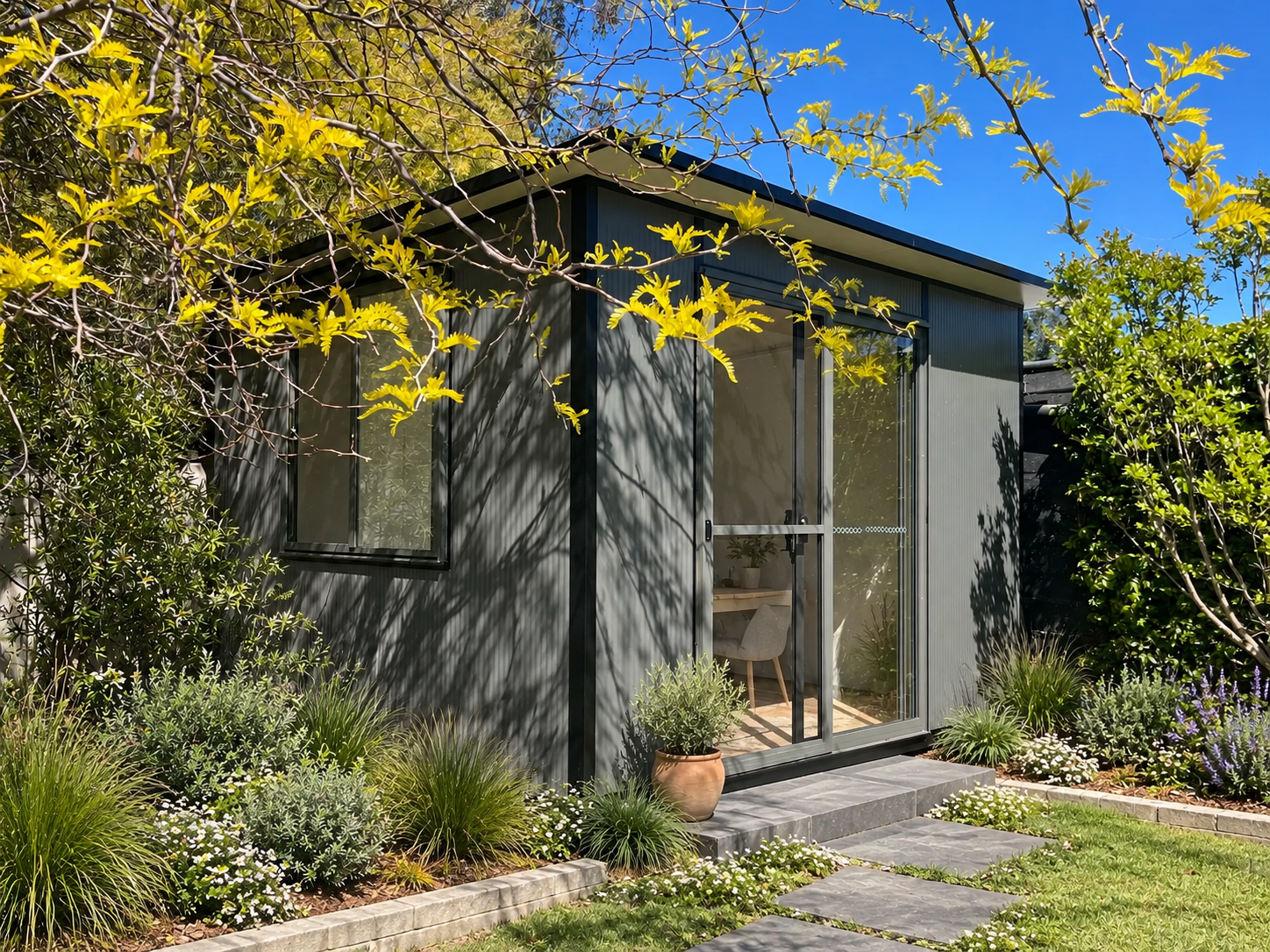 A modern gray Backyard Studio with large glass sliding doors and window, surrounded by lush green plants, shrubs, and flowering plants, with a stone pathway leading to the entrance, under a bright blue sky.