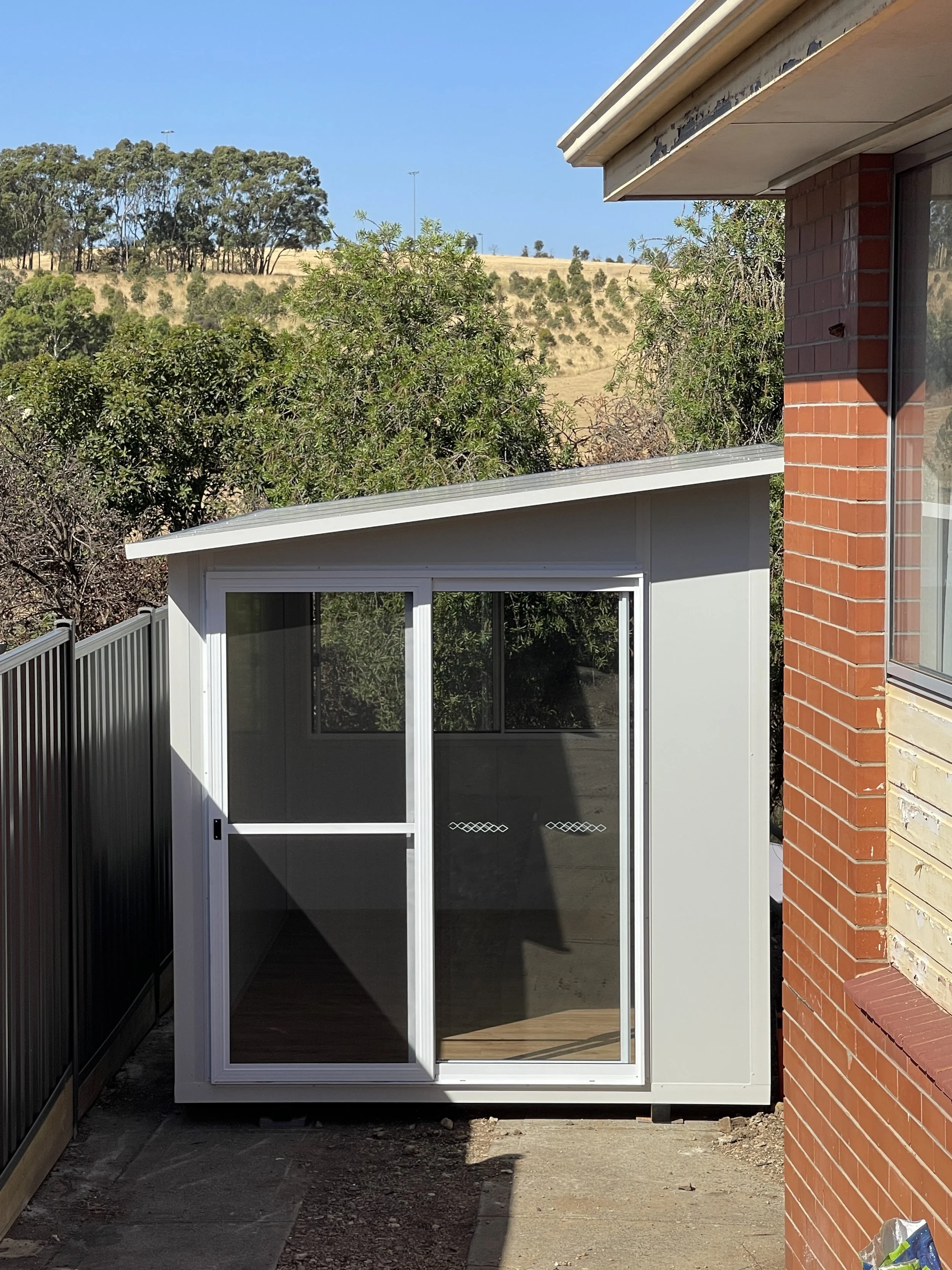 Small backyard pod with light grey cladding, sloped roof design, and sliding glass door, installed in a narrow side access area.