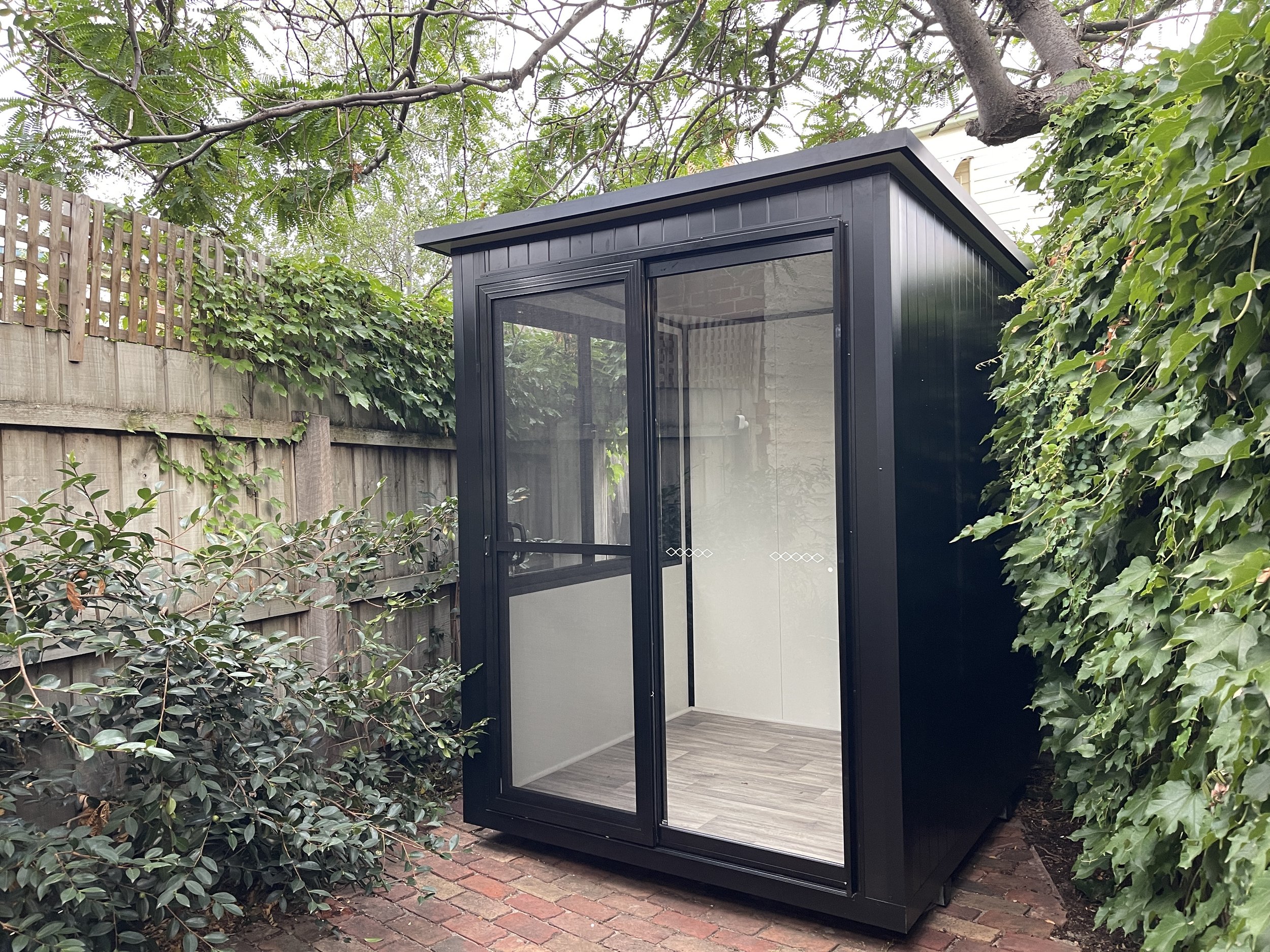 Modern black backyard office pod with large sliding glass doors, set on a brick patio and surrounded by greenery and wooden fencing.