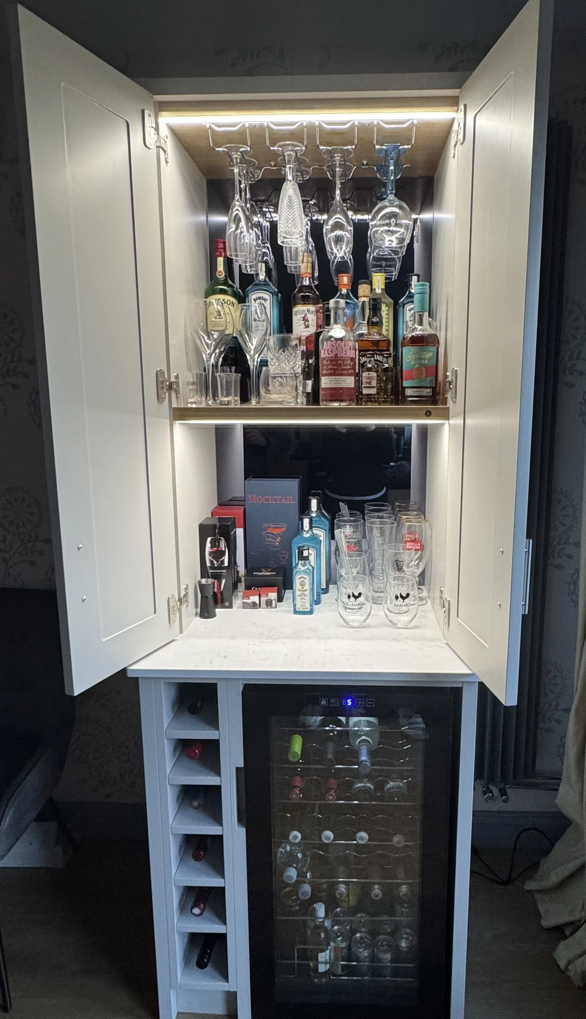 A home bar cabinet with open doors, containing liquor bottles, glassware, and a mini refrigerator with wine bottles inside. The cabinet has a wood top with LED lighting and a white finish, with wine bottle storage below.