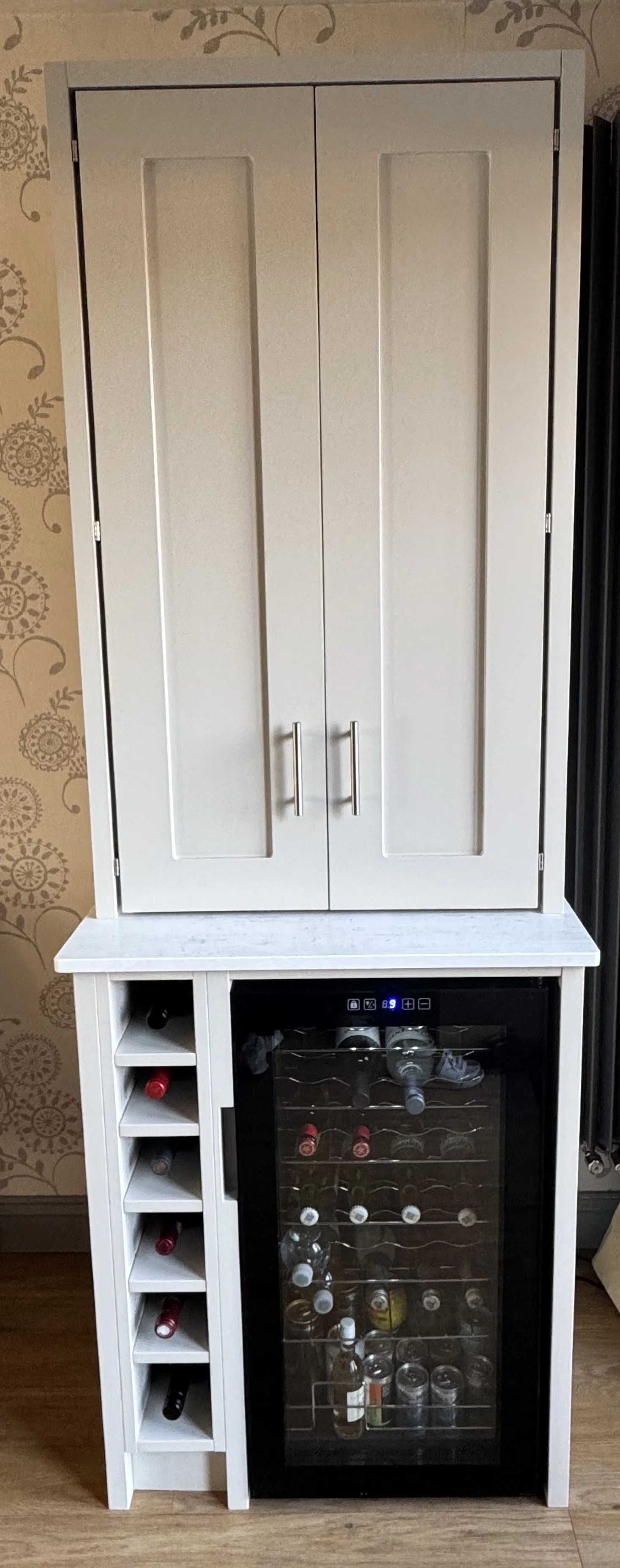 A white cabinet with double doors on top, a marble-like countertop, and a wine cooler with a glass door below. The wine cooler contains bottles, and to the left of it, there are slots with more wine bottles stored horizontally. The background has beige floral-patterned wallpaper.