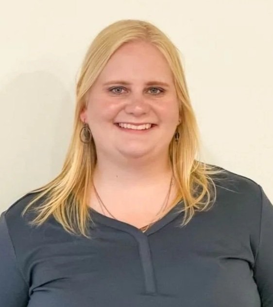 A woman with long blonde hair smiling at the camera, wearing a dark gray top and earrings, standing against a plain light-colored background.