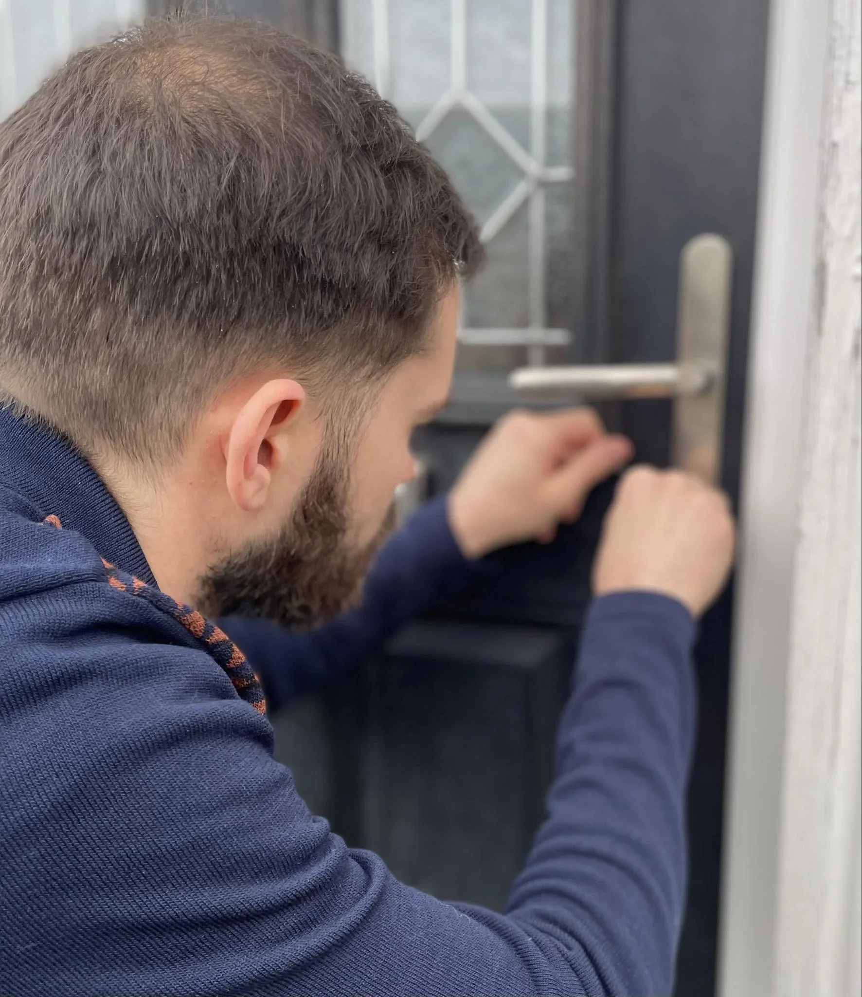 A man with a beard and short hair is kneeling by a door, working on the doorknob or lock.