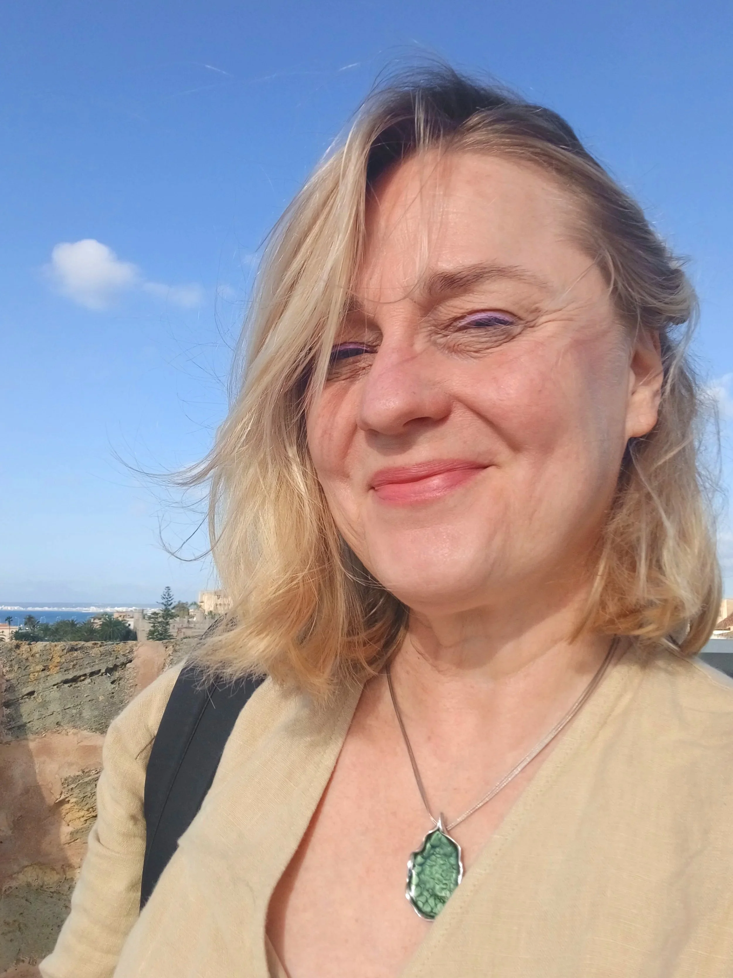 A smiling woman with blonde hair taking a selfie outdoors against a clear blue sky, with some clouds and a distant landscape in the background.