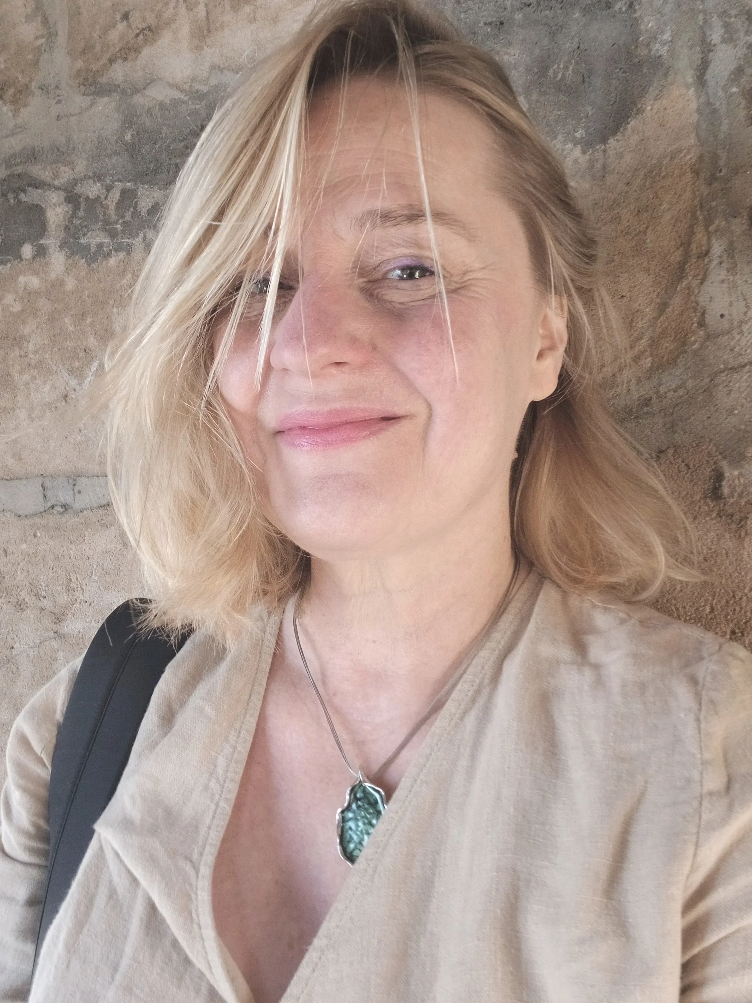 Close-up of a woman with blonde hair partially covering her face, wearing a beige shirt or jacket, a silver necklace with a green gemstone pendant, and a black strap, standing in front of a rustic stone wall.