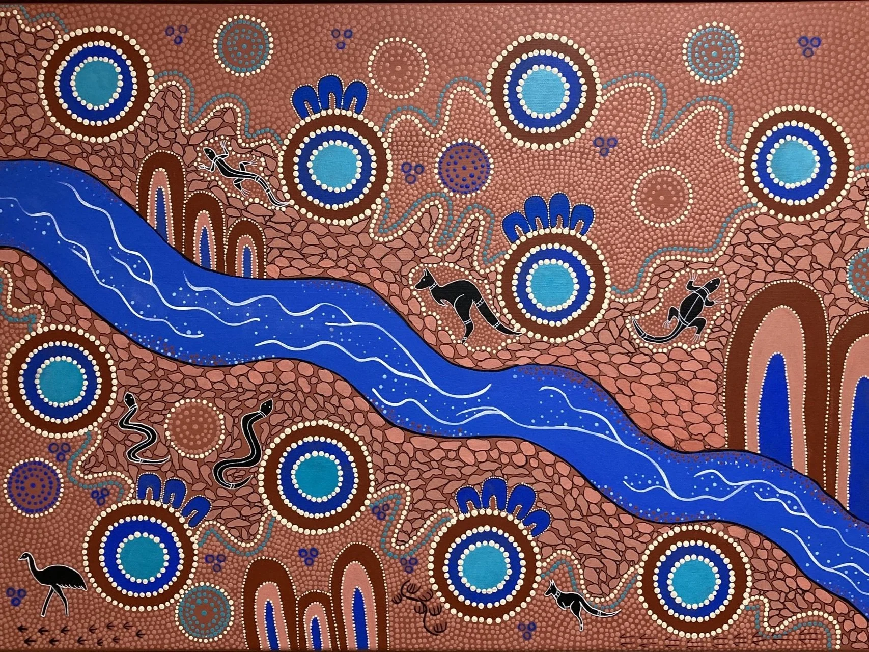 Aboriginal-style painting of a blue river flowing through a red-brown landscape with circles and dots. Black animals such as kangaroos, emus, and snakes are depicted among the scenery.