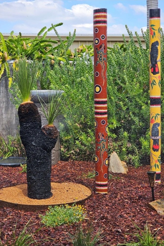 Decorative outdoor garden scene with three painted wooden poles and a black fountain sculpture resembling a cactus with two plants growing from its top.