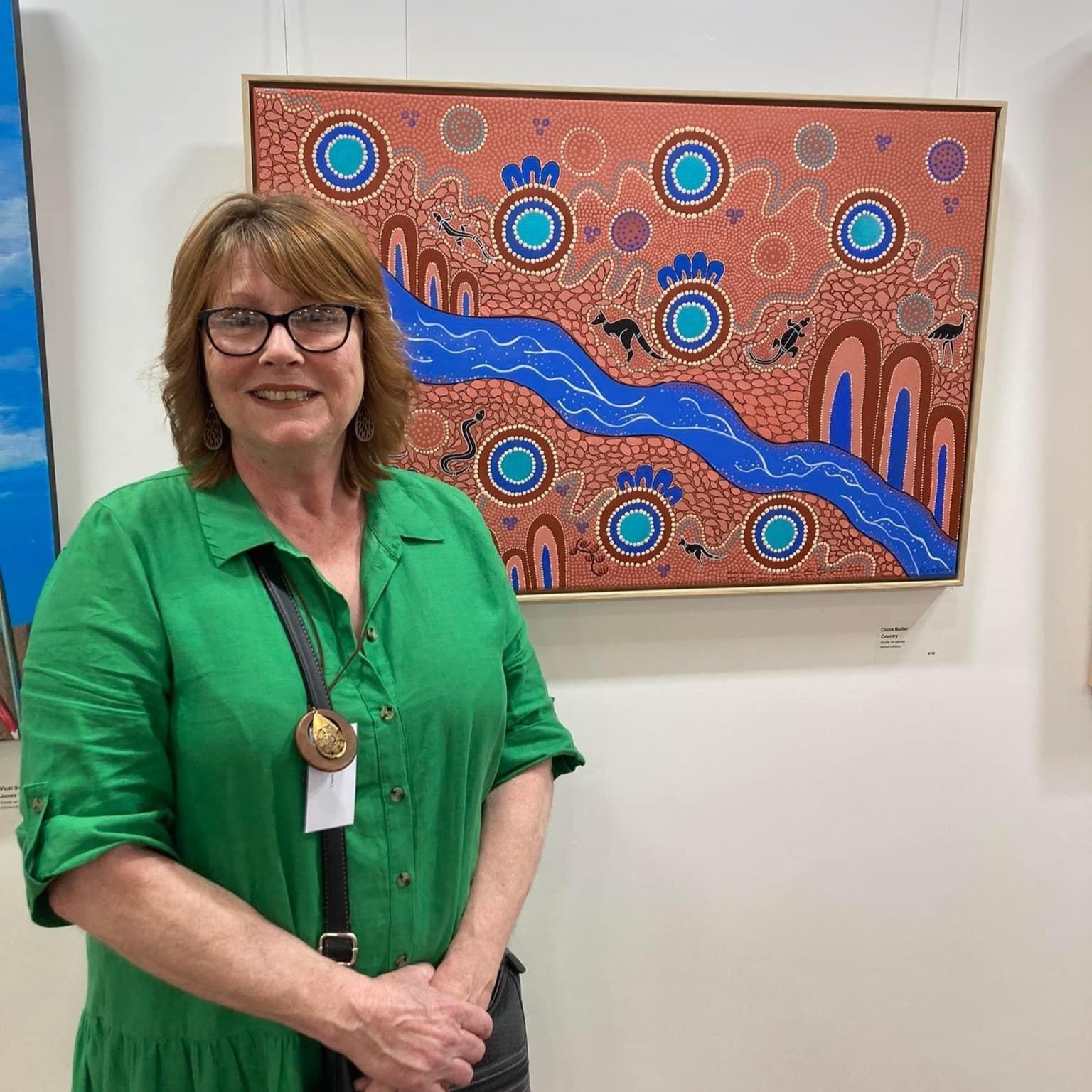 A woman with glasses, wearing a green dress, standing in front of an abstract painting with blue river-like lines, circles, and animal figures.