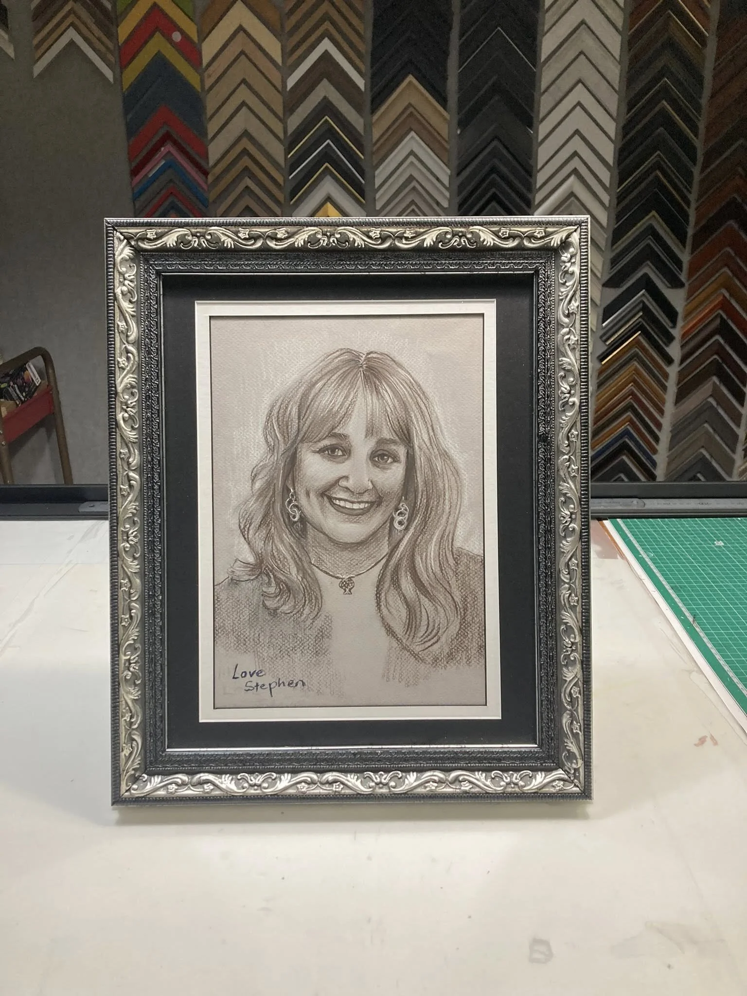 Framed pencil sketch of a woman with blonde hair, earrings, and a necklace, smiling. The drawing has a note at the bottom left reading 'Love Stephen.' Background shows a wall with a colorful zigzag pattern.
