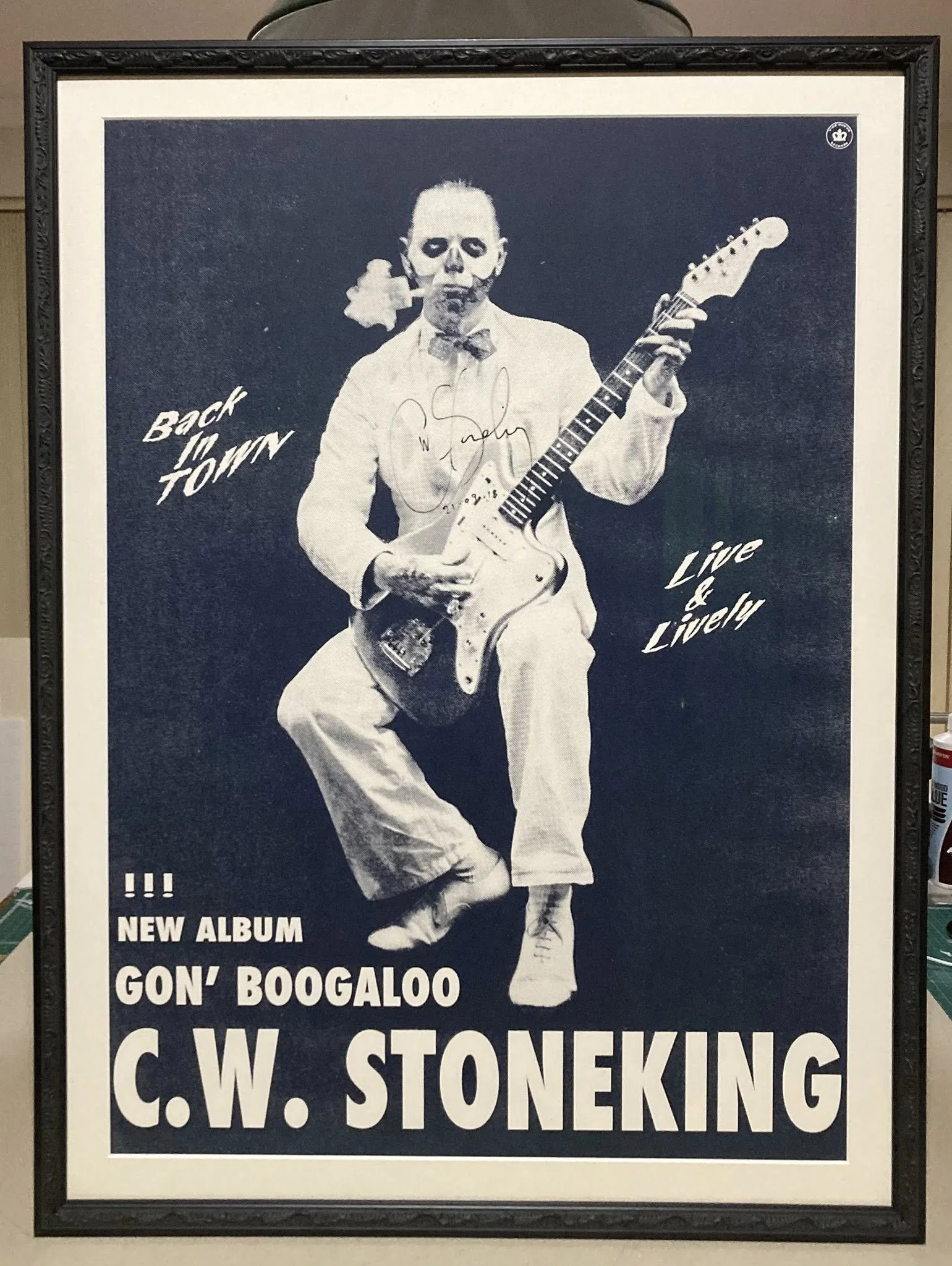 Black and white poster of a person in spooky makeup playing an electric guitar. The poster promotes C.W. Stoneking's new album 'Gon' Boogaloo' with phrases 'Back in Town' and 'Live & Lively' and includes an autograph from the artist.