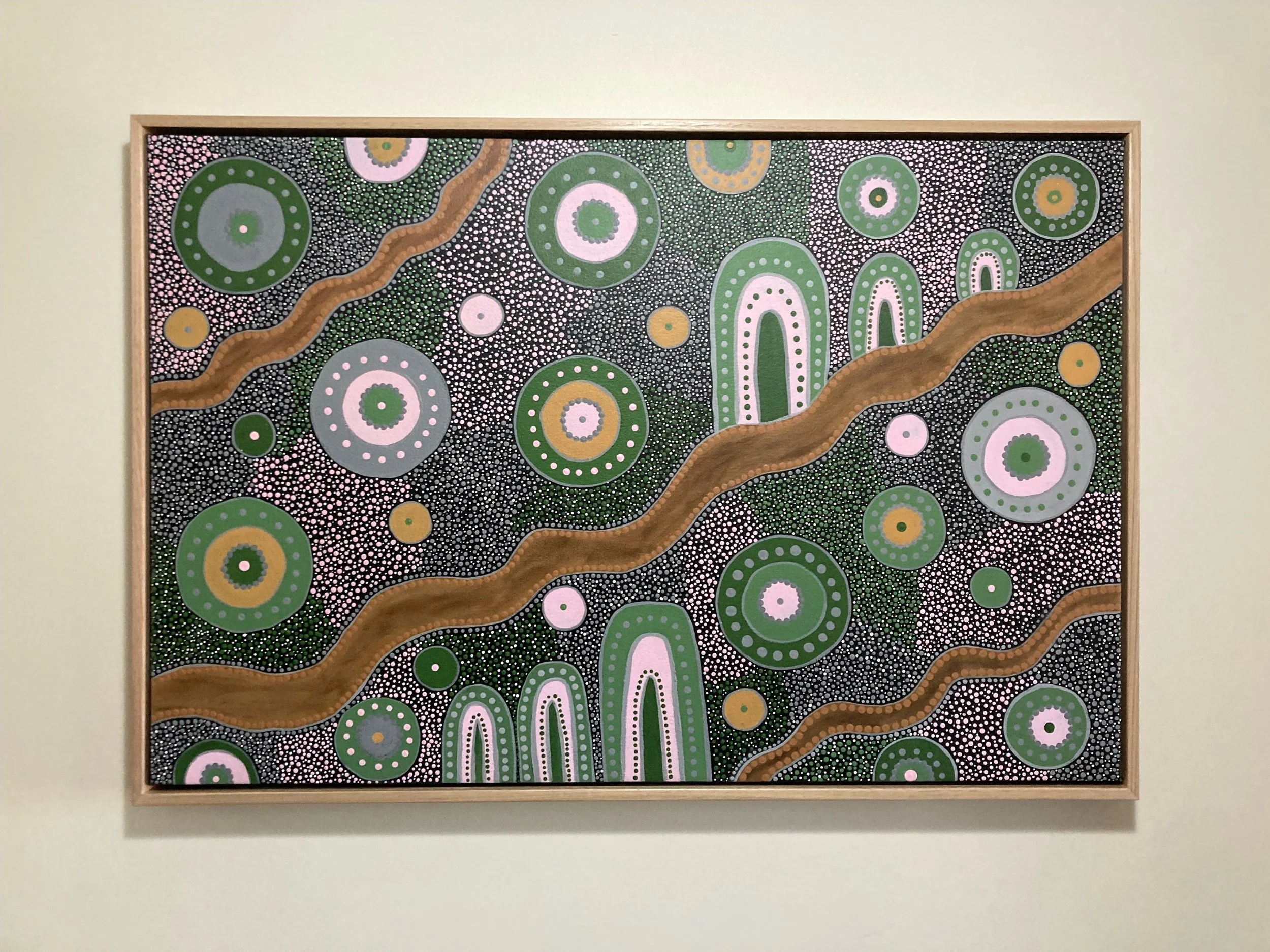 An abstract painting with green and pink circular patterns, white dots, and two brown wavy lines on a black background, framed in light wood.
