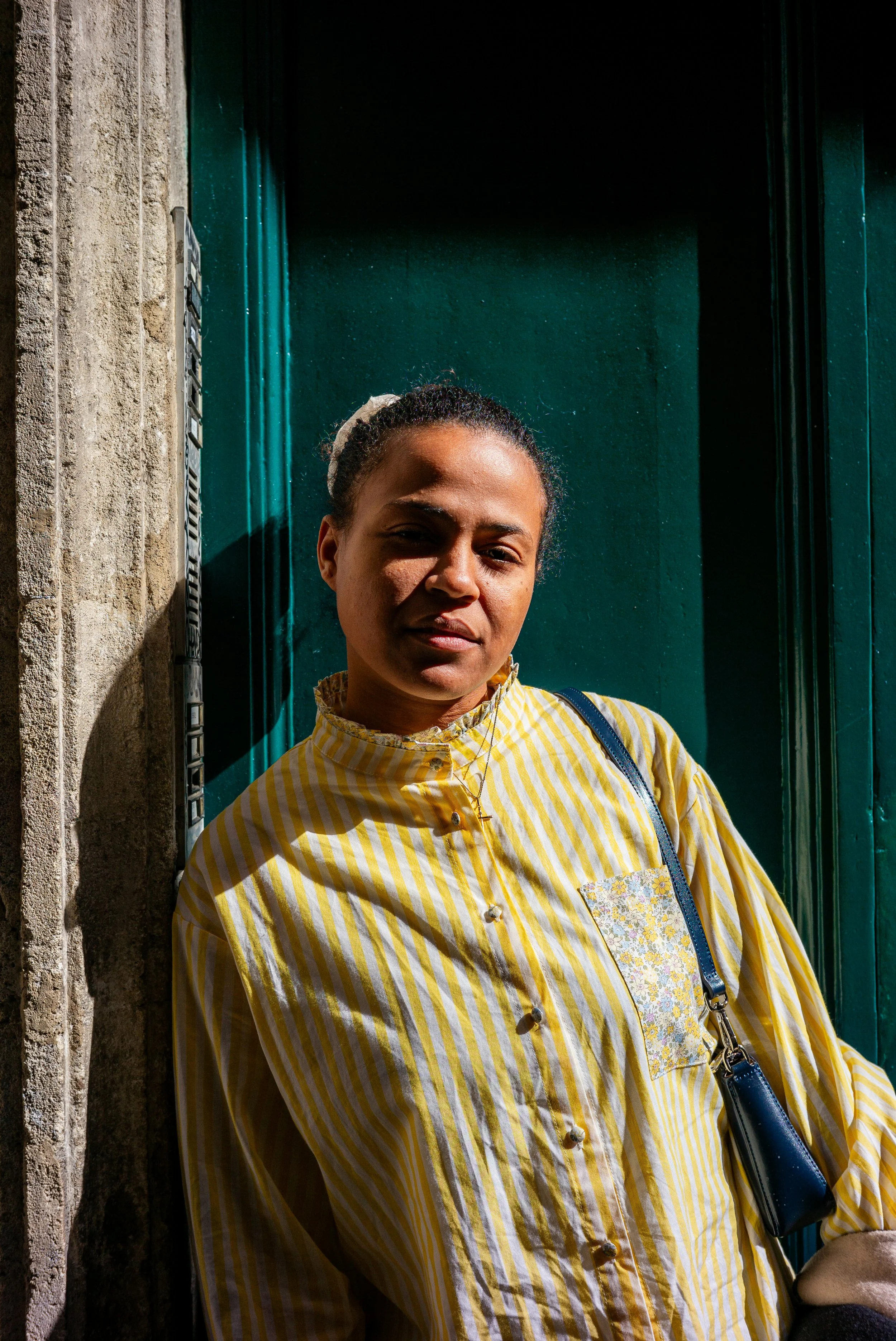 Jeune femme souriante portant un chemisier à rayures jaunes et blanches, avec un sac à l'épaule, debout devant une porte verte, ensoleillement contrasté.