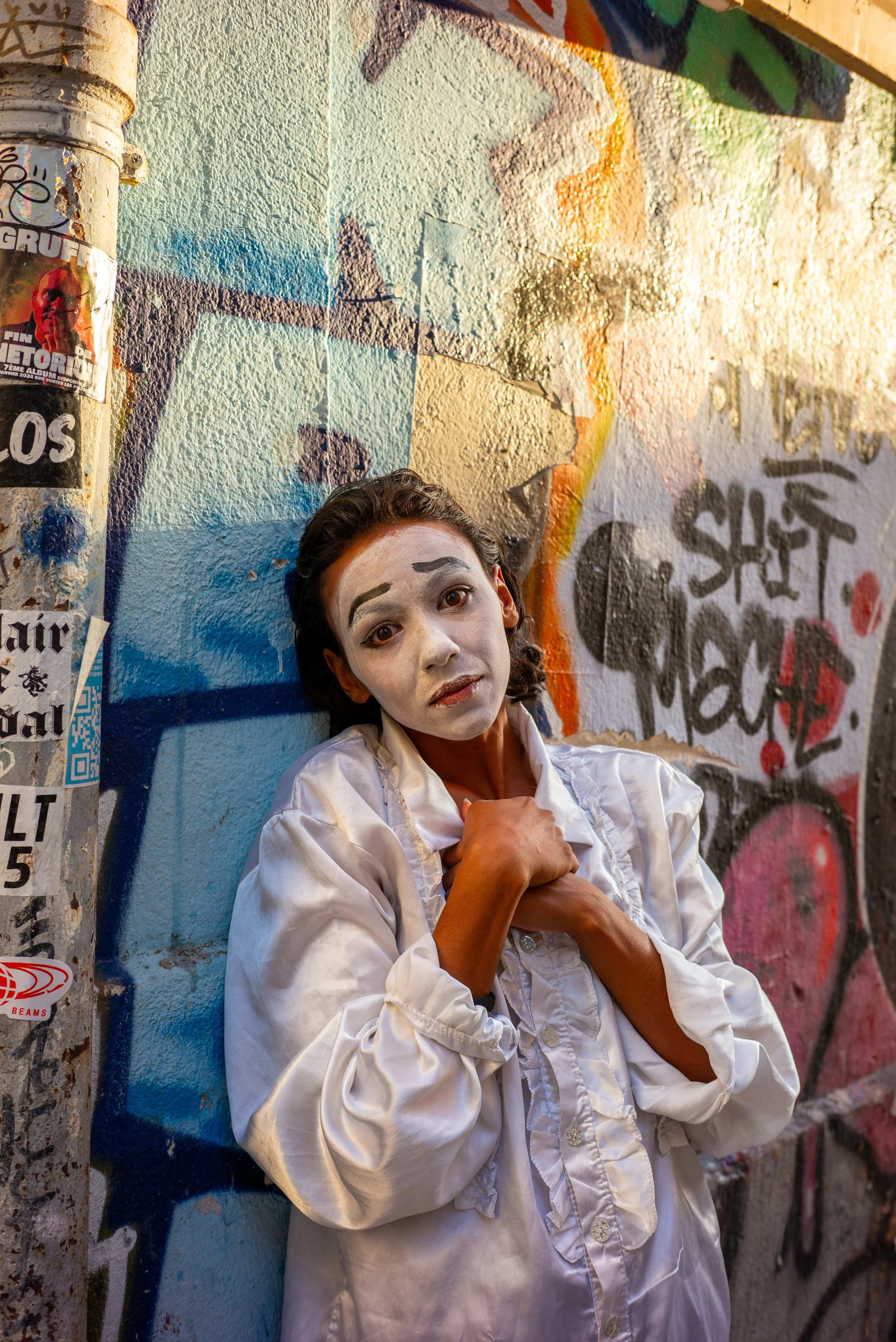 Une femme avec un visage maquillé en blanc, portant une veste blanche, est appuyée contre un mur couvert de graffitis colorés.