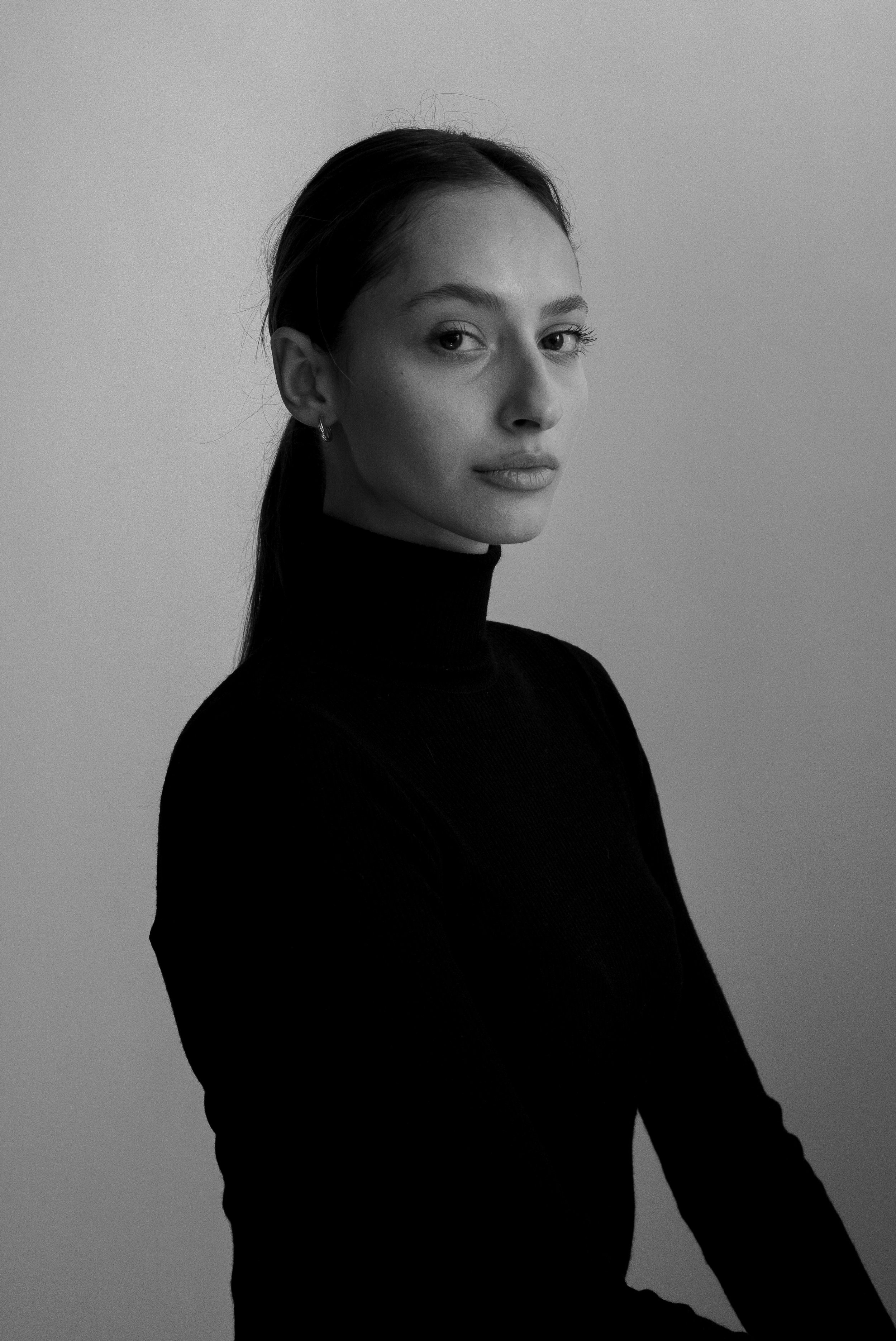 Portrait en noir et blanc d'une femme avec pull noir, regardant vers la caméra, fond gris clair.