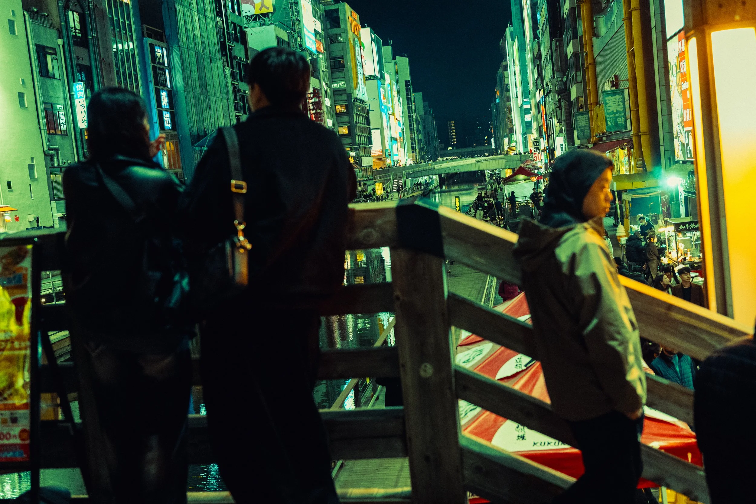 Trois personnes, deux femmes et un enfant, regardant sur une passerelle dans un quartier animé la nuit, avec des enseignes lumineuses en japonais sur des bâtiments modernes.