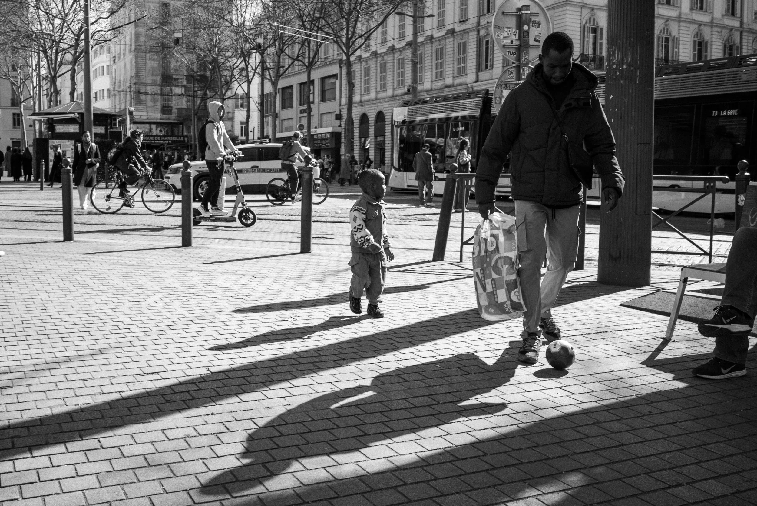 Une scène urbaine en noir et blanc avec des personnes marchant, faisant du vélo et jouant au foot, sous un ciel ensoleillé.