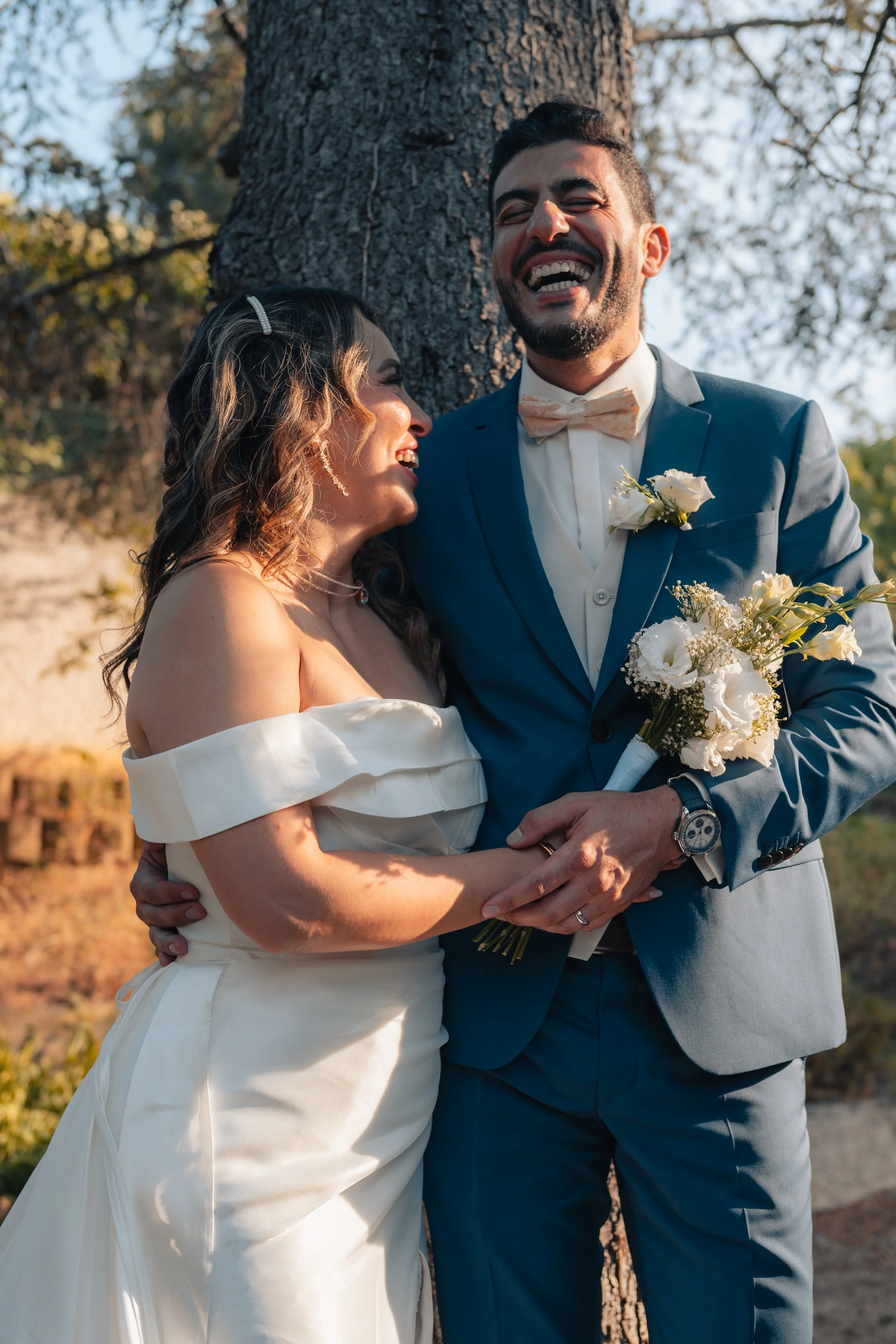 Un couple de mariés qui sourient et s'embrassent devant un arbre en plein air.