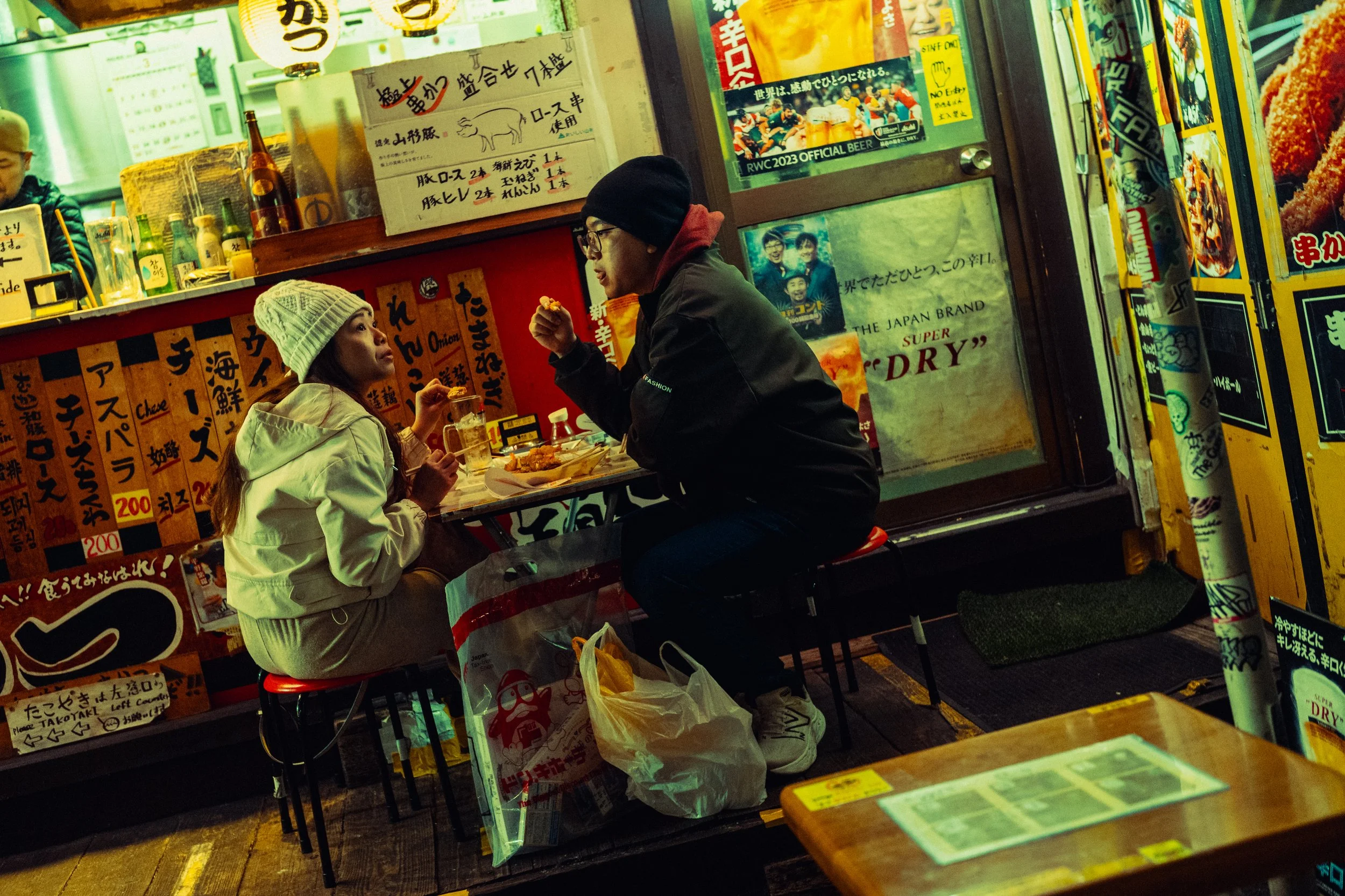 Deux personnes mangent dans un restaurant japonais de nuit, assises face à face, avec des affiches et des enseignes en japonais autour, dans une ambiance colorée et animée.