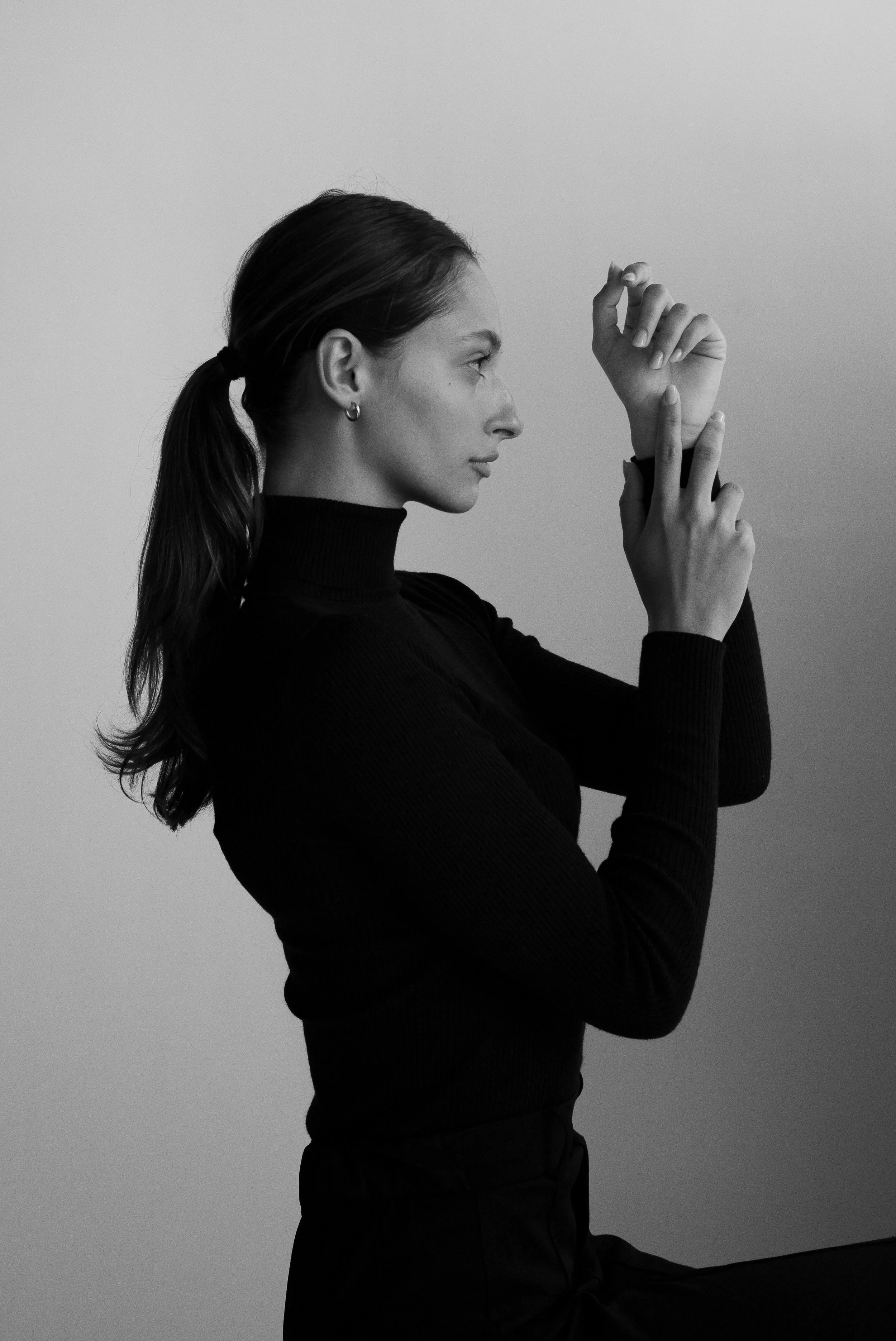 Femme en noir et blanc, vue de profil, portant un pull à col roulé, avec les cheveux attachés en queue de cheval, levant une main près de son visage.