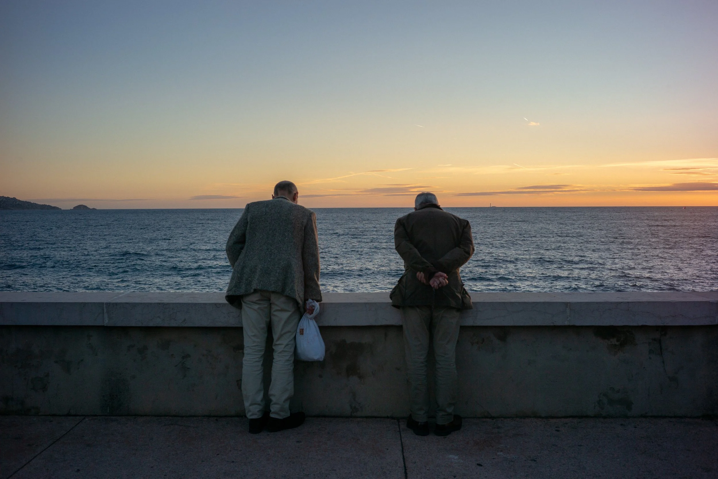 Deux hommes regardent la mer au coucher du soleil, un à gauche avec un manteau gris et un sac blanc, l'autre à droite avec un manteau noir, tous deux de dos.