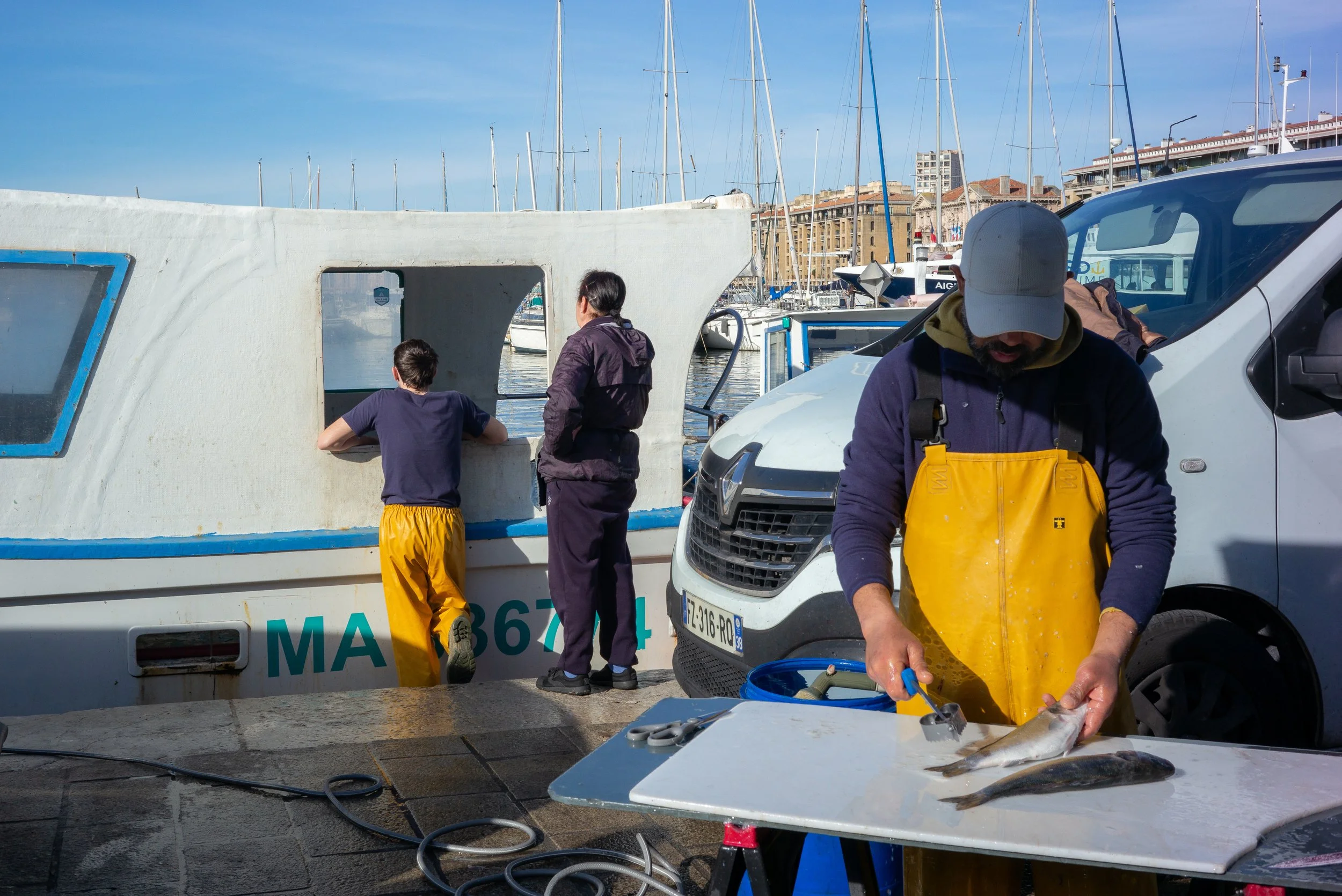 Des pêcheurs nettoient des poissons sur une table près d'un port avec des bateaux à voile en arrière-plan, par beau temps ensoleillé.