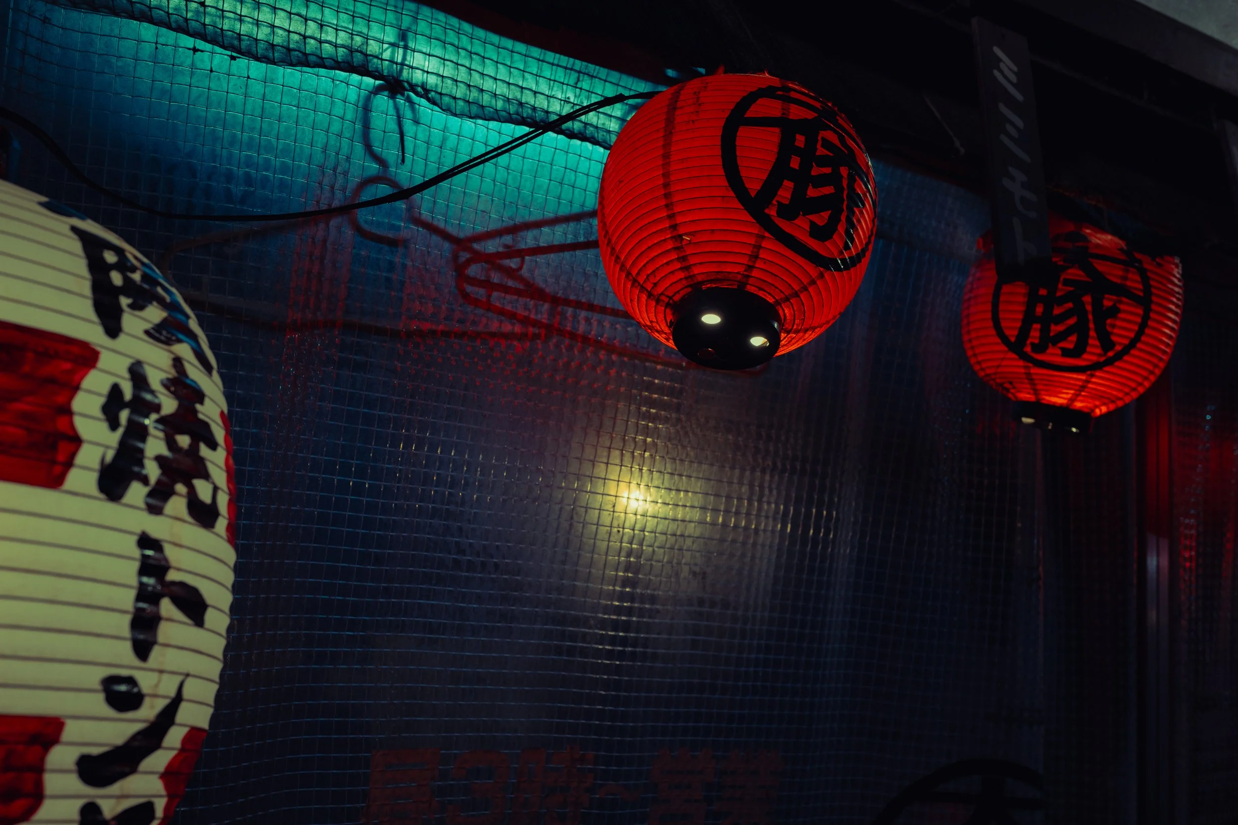 Lanterne japonaises rouges et une lanterne blanche suspendues à un plafond noir, éclairées et reflétant une lumière chaude, avec un fond de grille métallique bleue.