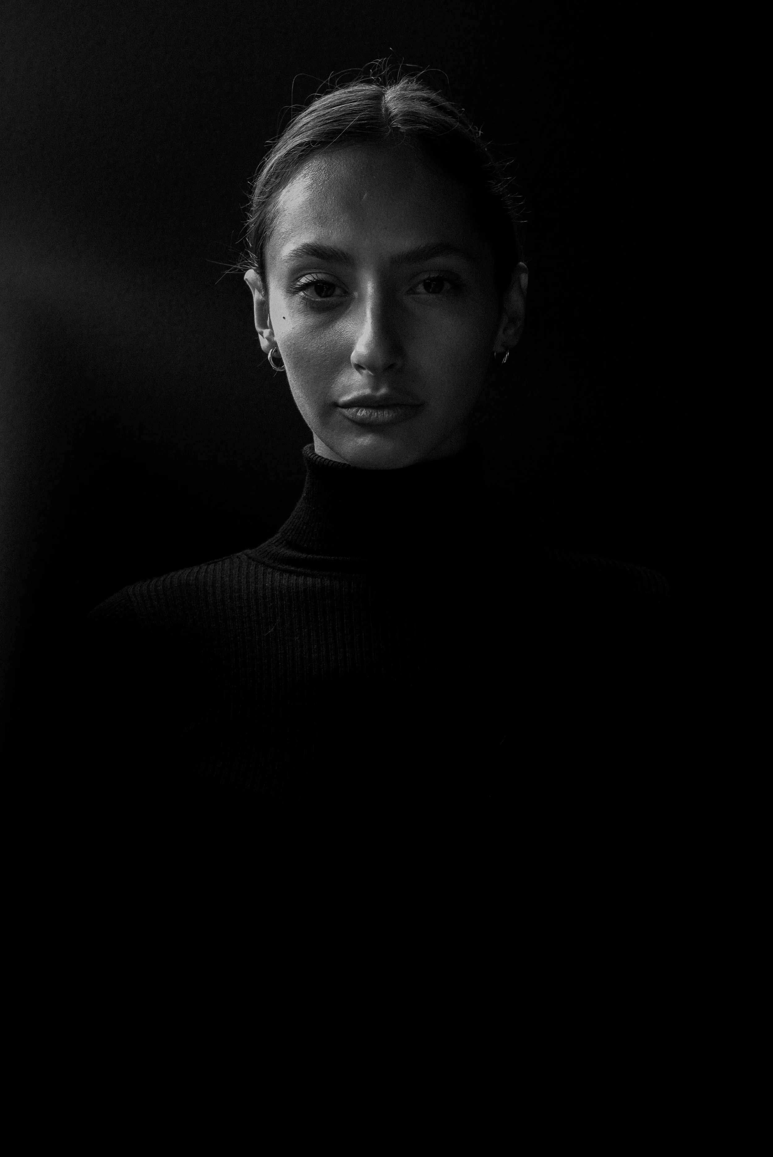 Portrait en noir et blanc d'une femme avec un pull moulant, cheveux attachés, et expression calme, sur un fond sombre.