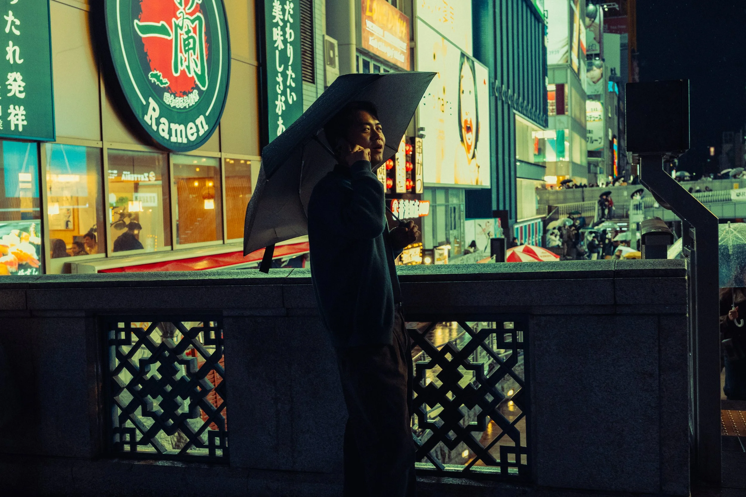 Un homme se tient sur un pont ou une rampe, tenant un parapluie, dans une rue animée de nuit, avec beaucoup de lumières colorées et des enseignes en japonais, certains montrant des restaurants et des commerces.