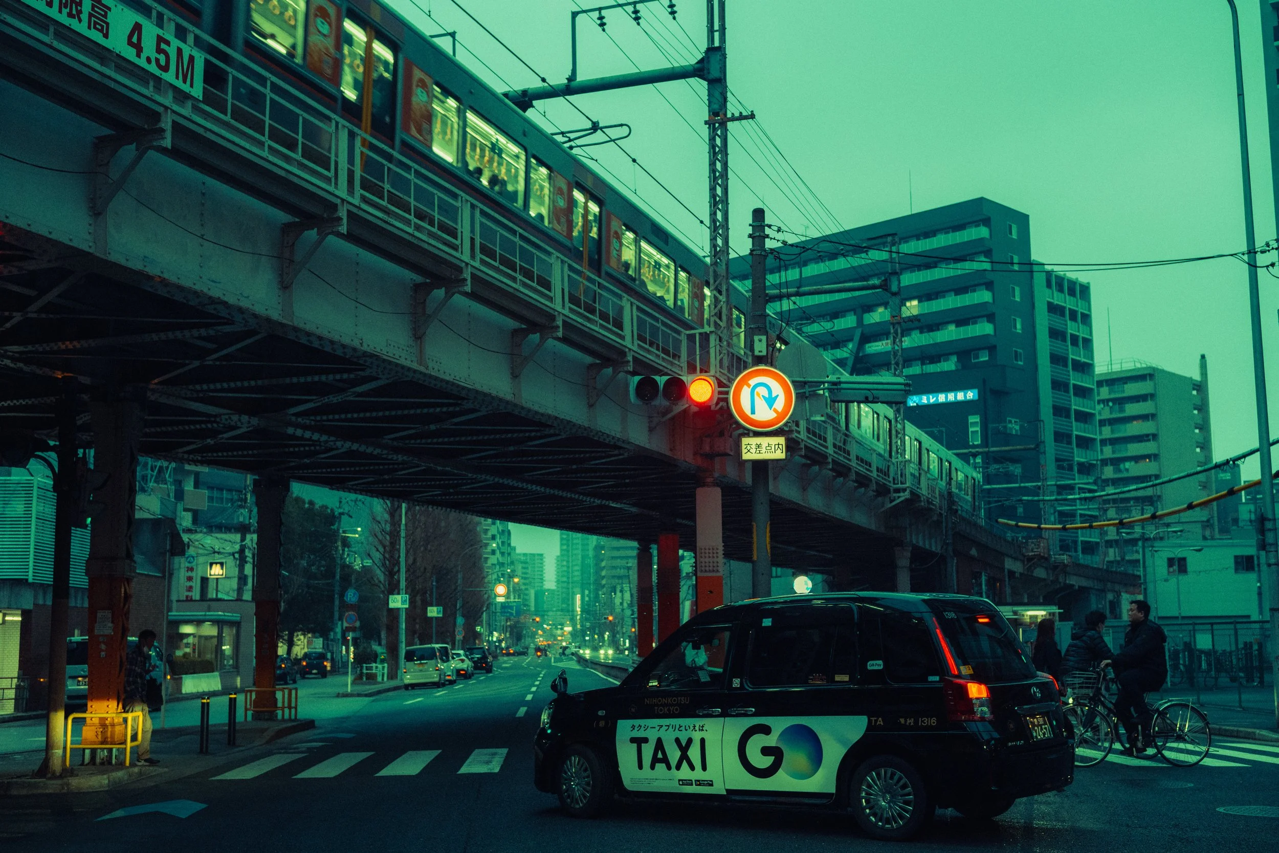 Une scène urbaine avec un métro passant au-dessus d'une rue et un taxi électrique en bas, avec des piétons et des cyclistes. La scène est dans une ville japonaise, avec des panneaux de signalisation en japonais et des bâtiments modernes en arrière-pl