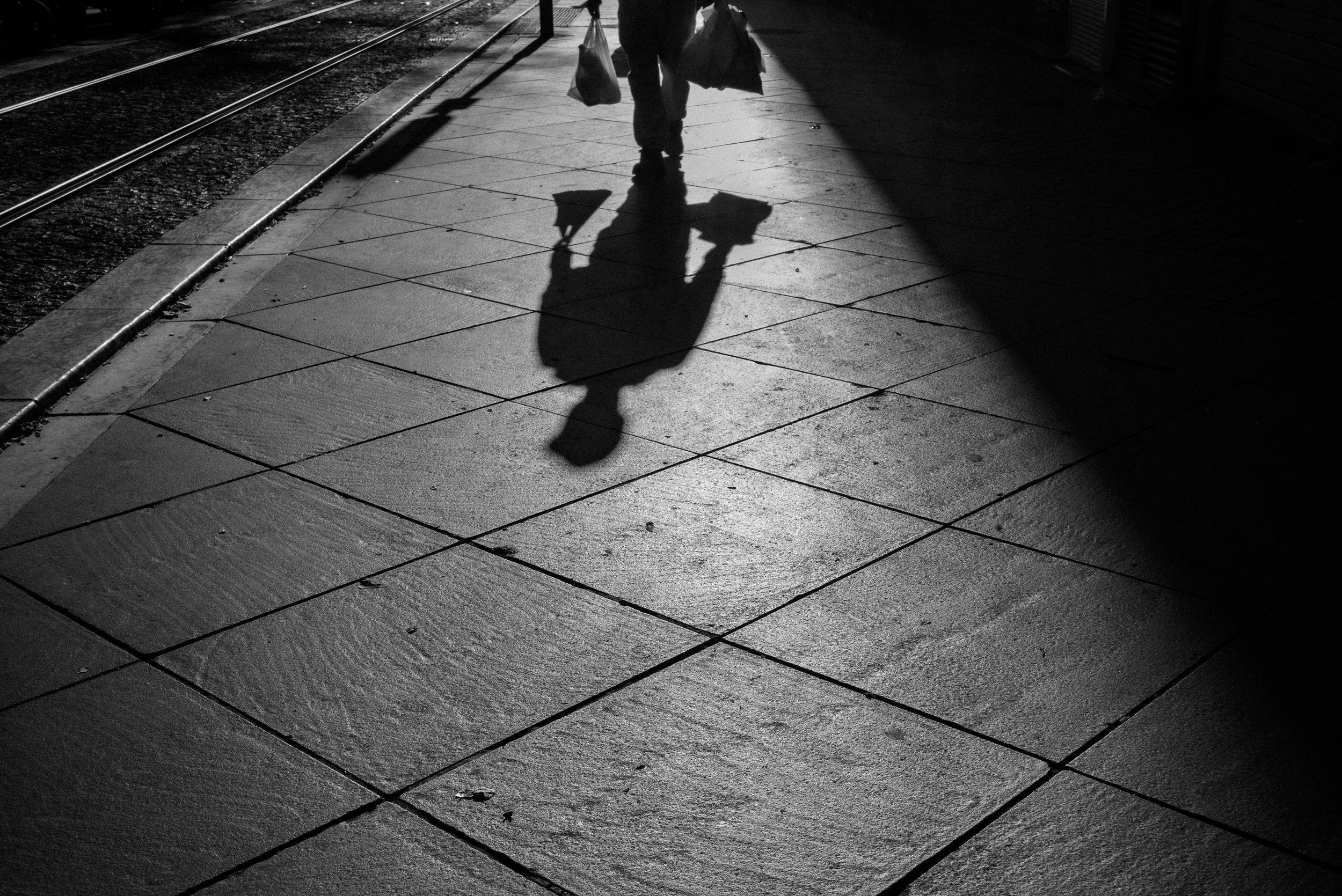 Une silhouette d'une personne portant deux sacs marche sur un trottoir ensoleillé lors de la fin du jour ou début de la nuit, créant une ombre longue.
